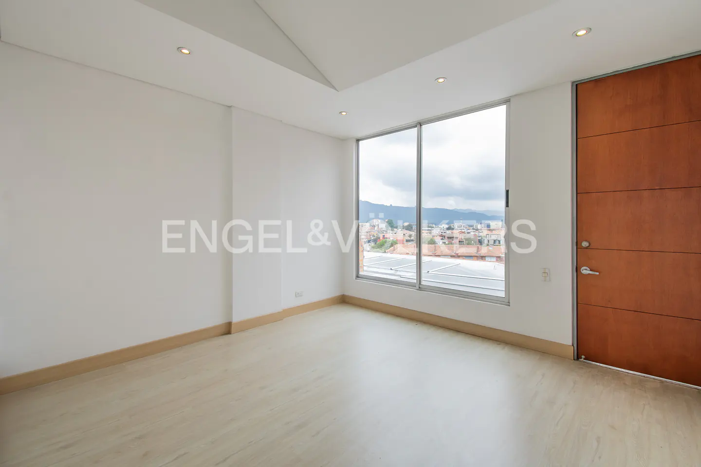 Bright, empty room with light wood floors, white walls, and a large window with a city view. A wooden door is on the right.