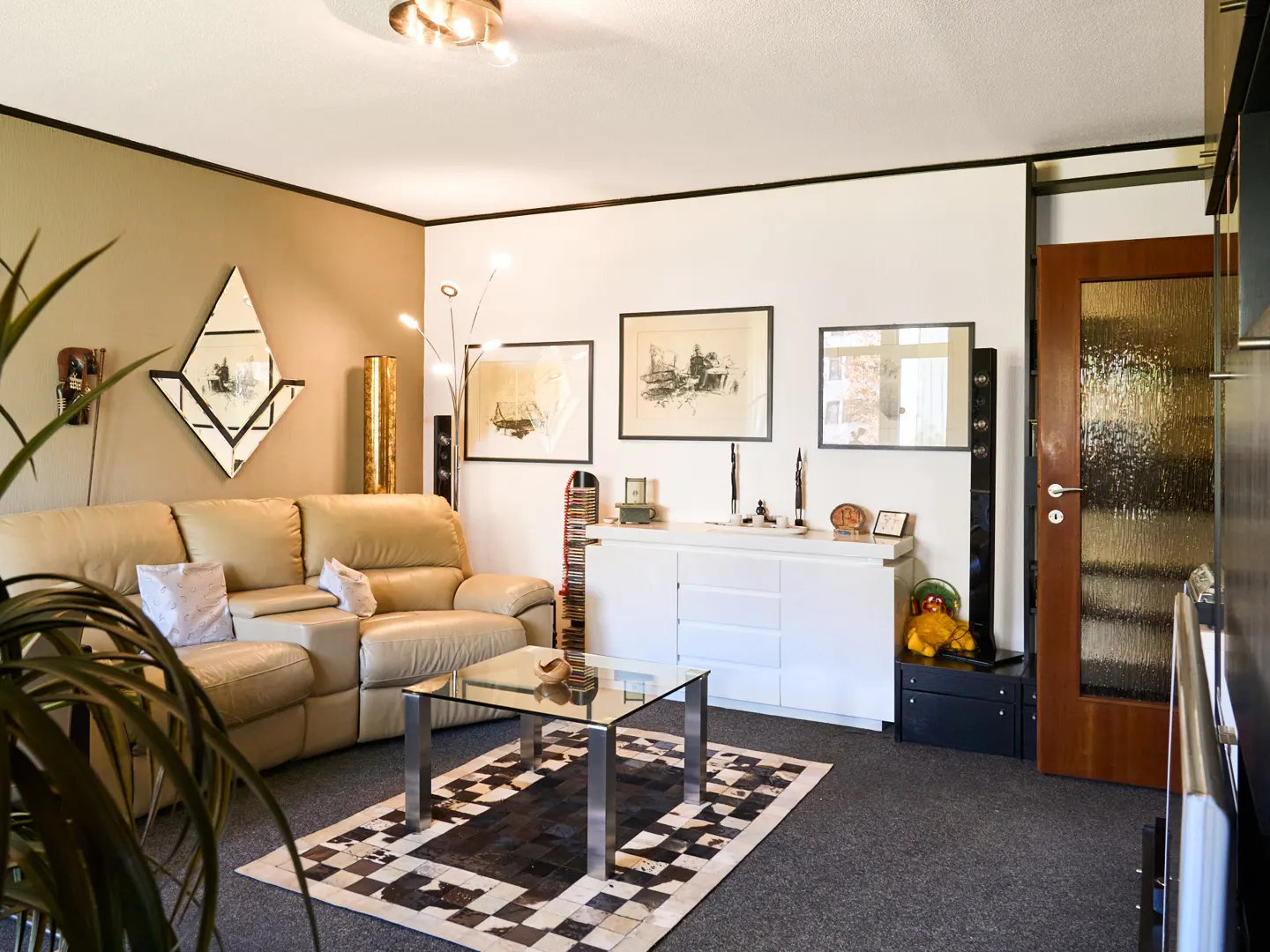 Living room with beige leather sofa, glass coffee table on a patterned rug, and white sideboard with framed art above.