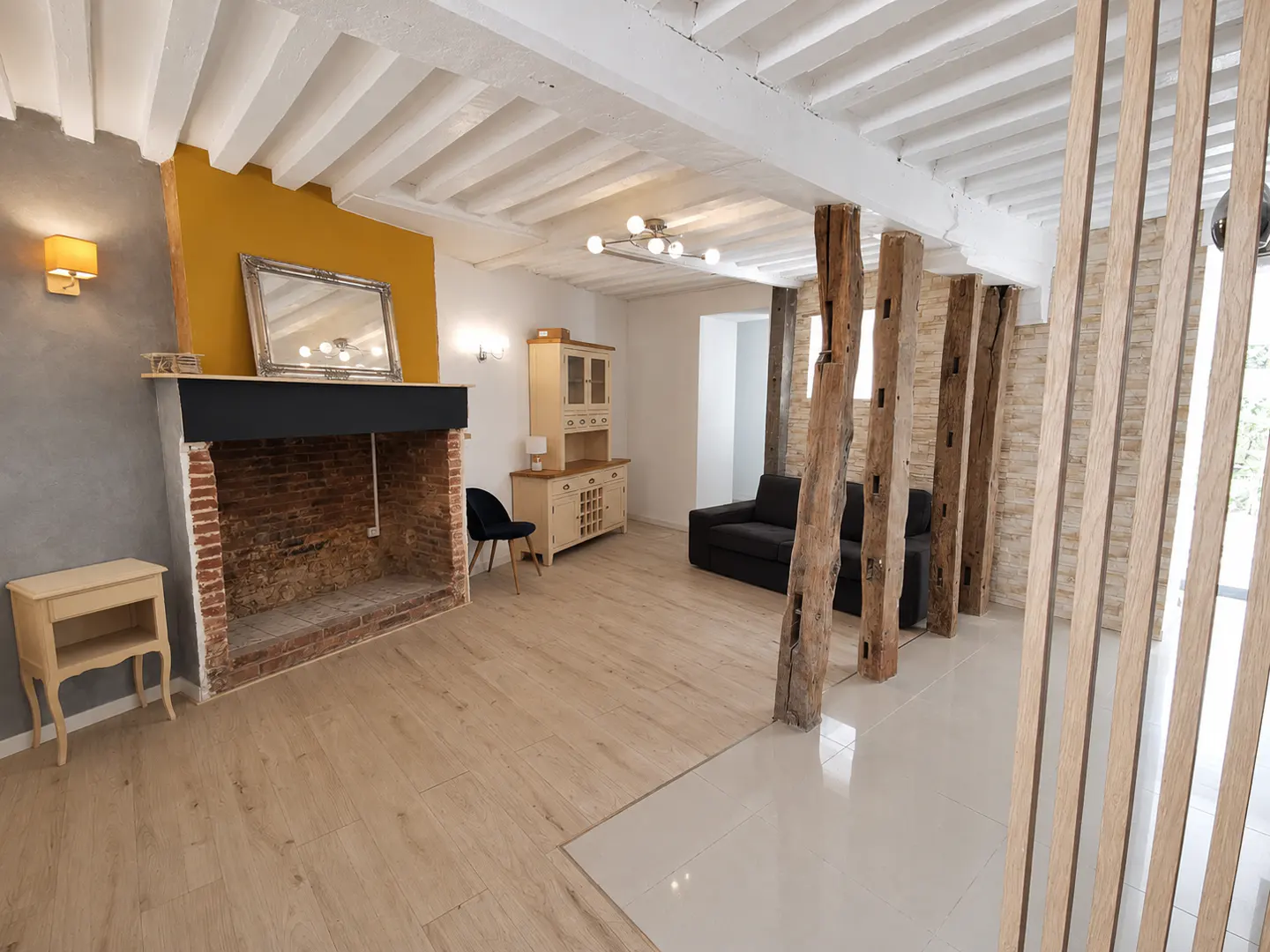Living room with exposed wooden beams, brick fireplace, mustard accent wall, and a black sofa in a rustic-modern home.