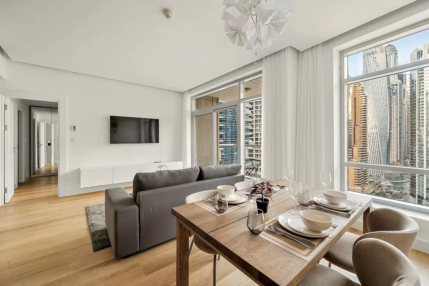 Bright apartment interior with wood floors, a gray sofa, and a dining table set for four. Large windows offer a city view.
