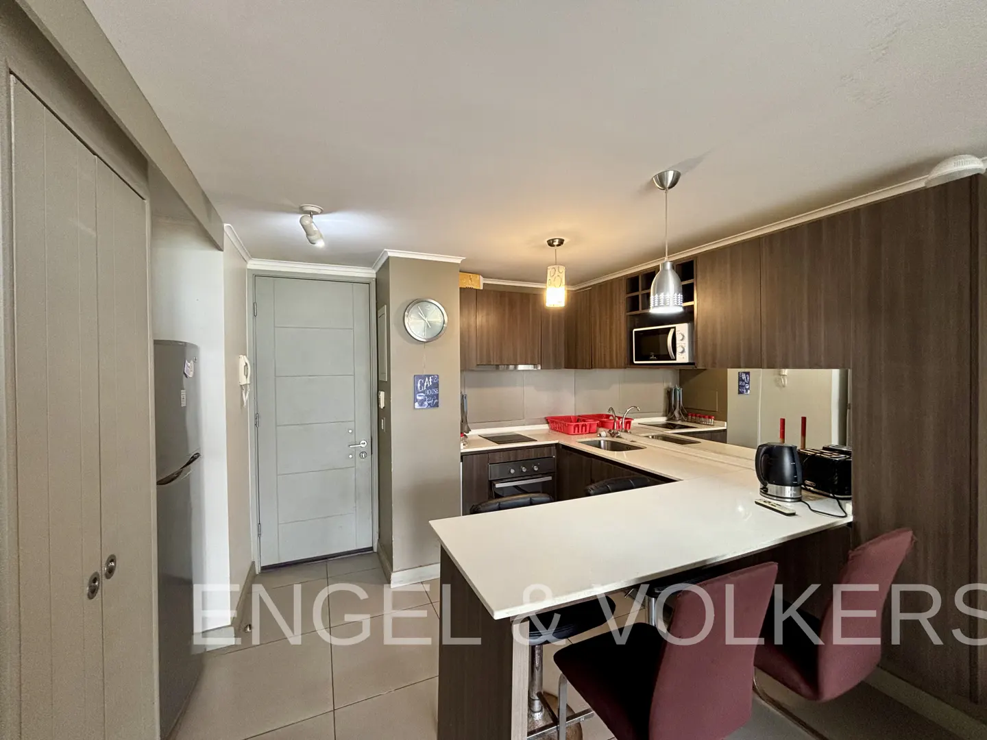 A modern kitchen with dark wood cabinets, a white countertop island with two red bar stools, and stainless steel appliances.