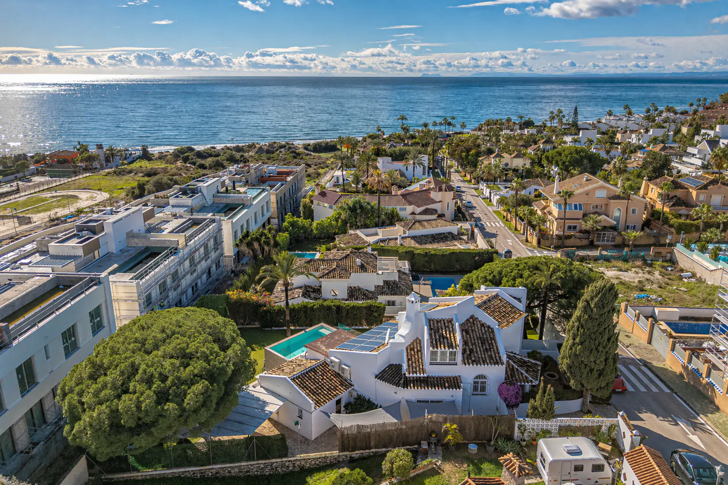 Aerial view of a coastal neighborhood with white houses, green trees, and a blue ocean under a partly cloudy sky.
