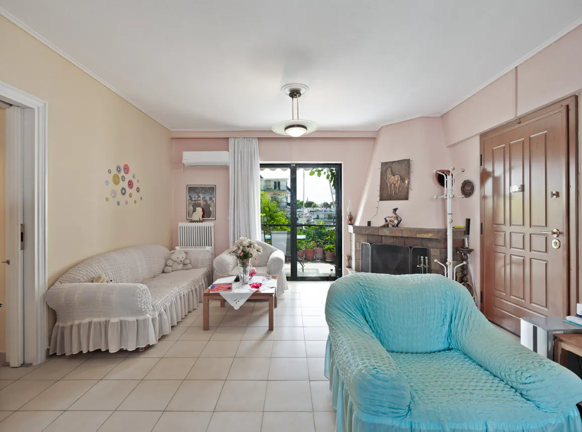 Living room with a light blue armchair, white sofa, fireplace, and a balcony with greenery. The walls are painted in light pink and beige.