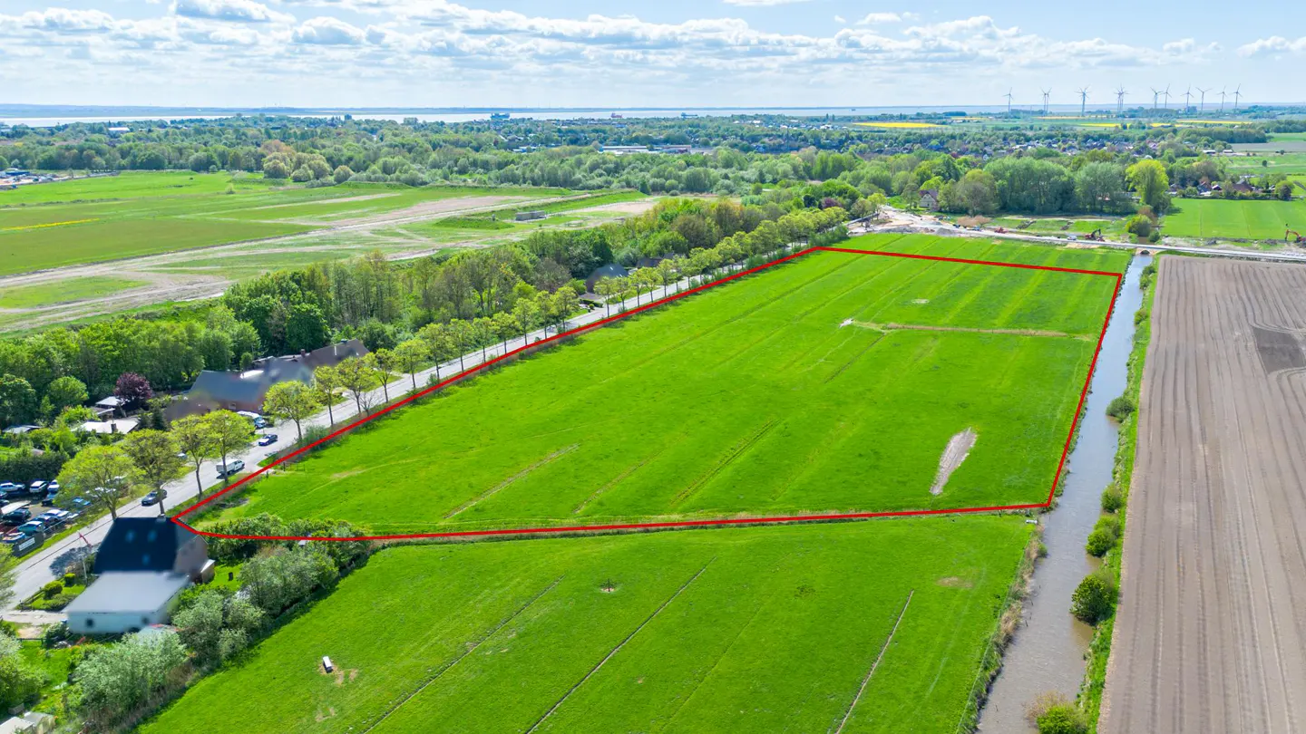 Aerial view of a large, rectangular green field outlined in red, bordered by trees and a road.