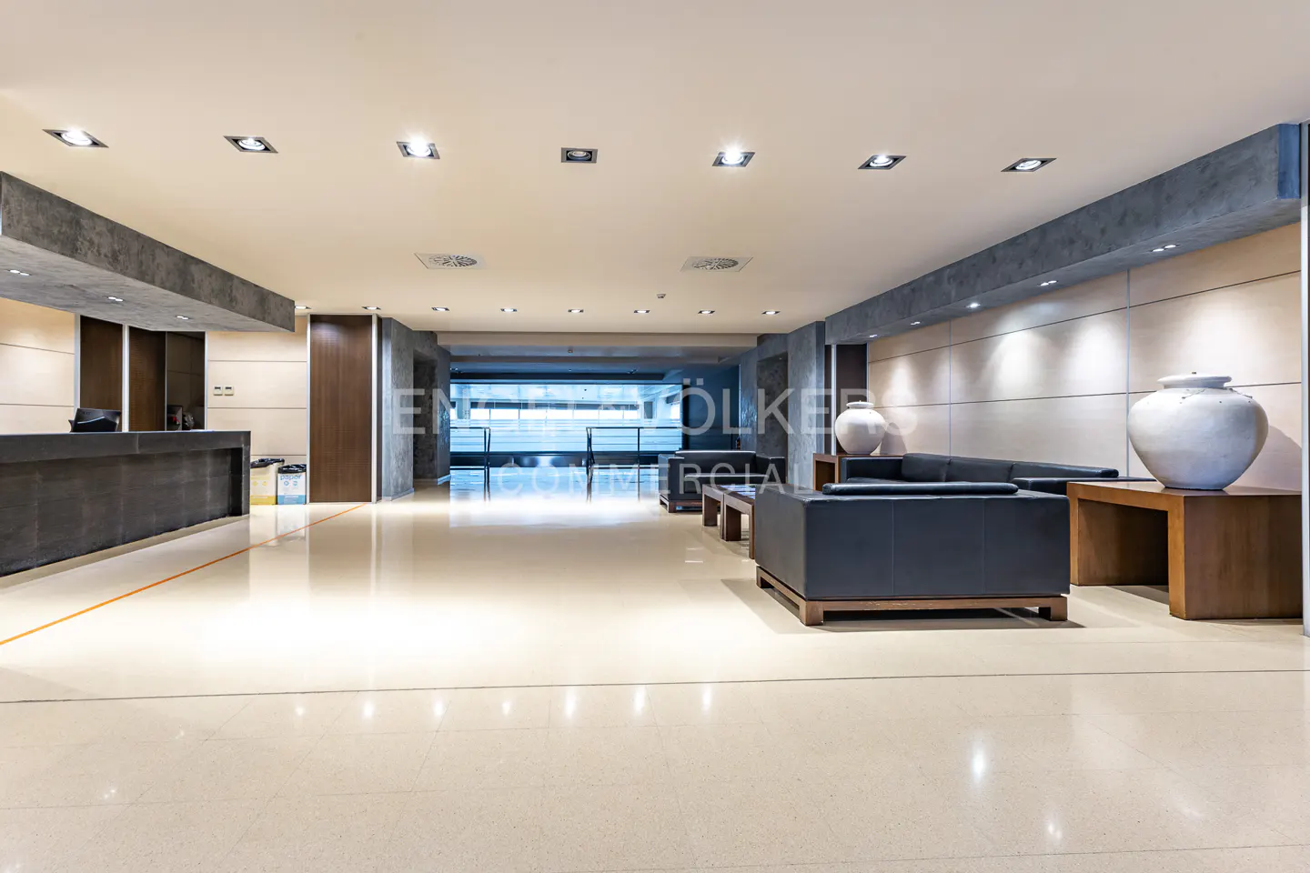 Hotel lobby with a reception desk, seating area with a black sofa, and large white vases on wooden tables. The floor is beige and shiny.
