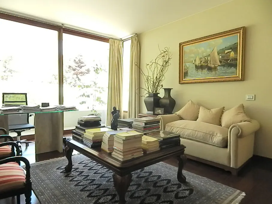 Living room with a large window, desk, sofa, and a coffee table piled with books. A painting hangs above the sofa.