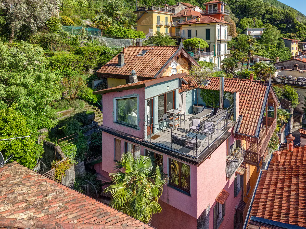 A pink house with a rooftop terrace featuring outdoor furniture, nestled in a lush, hilly Italian village setting.