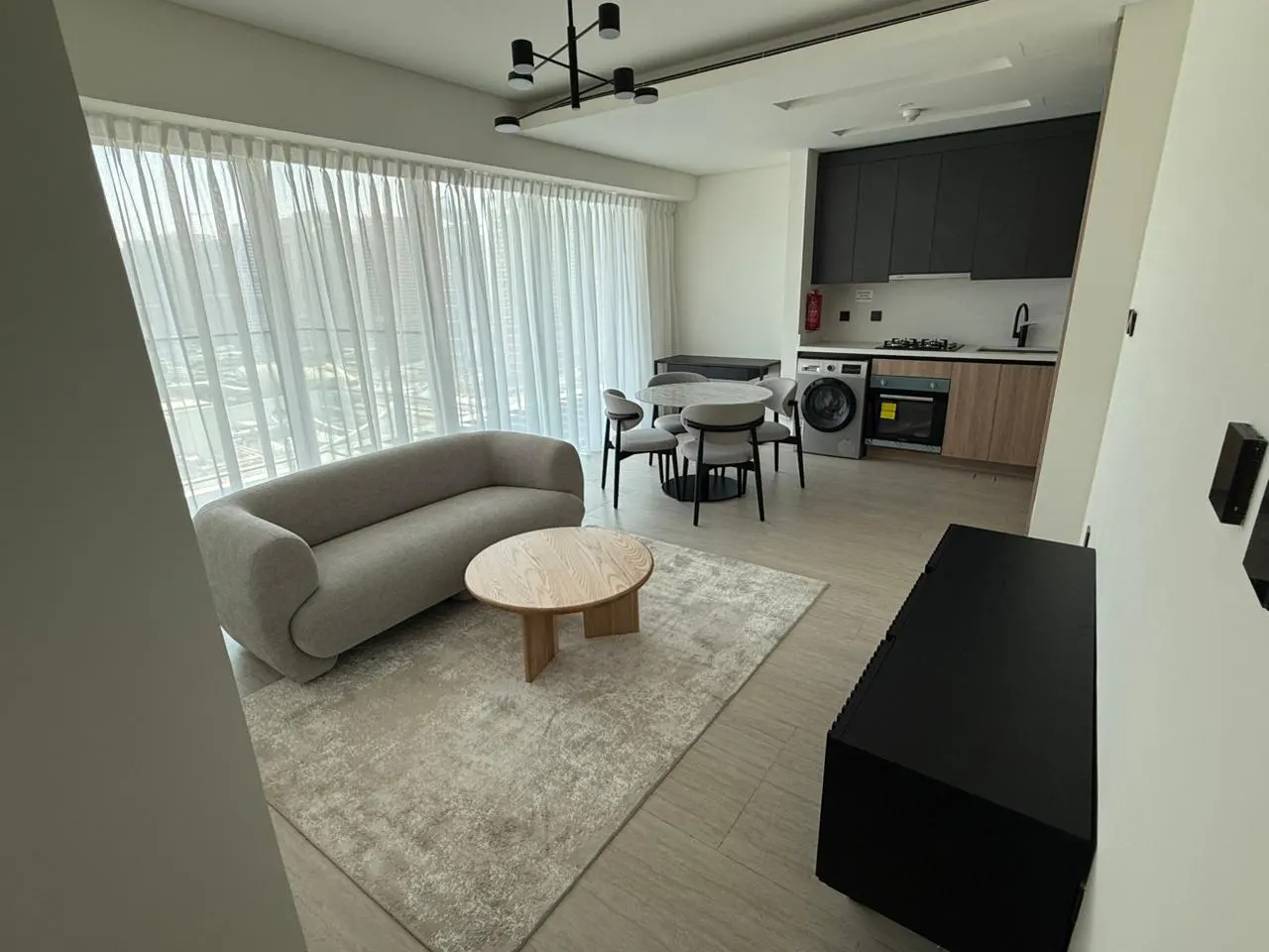 Modern open-plan apartment with a grey sofa, round wooden coffee table, dining area, and kitchen with dark cabinets.