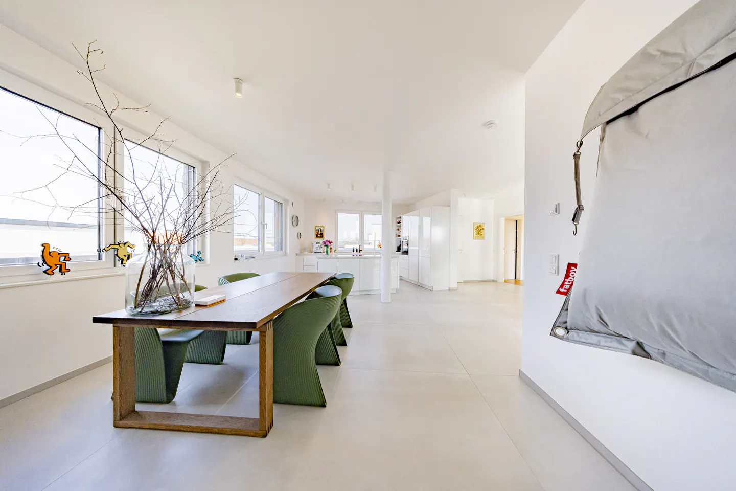 Bright, modern open-concept home with a wooden dining table, green chairs, and a white kitchen. A large gray Fatboy beanbag hangs on the wall.