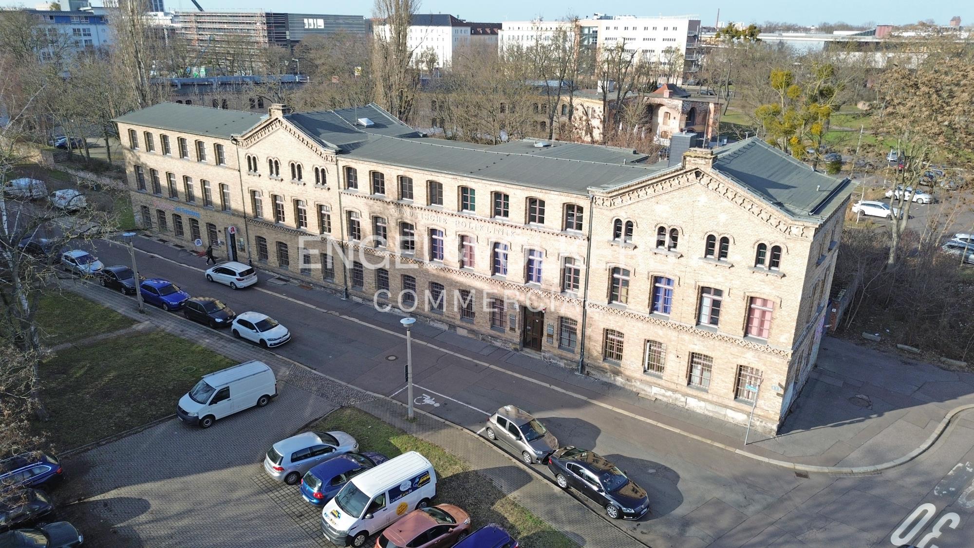 Aerial view of a long, multi-story brick building with a grey roof, situated next to a street with parked cars.