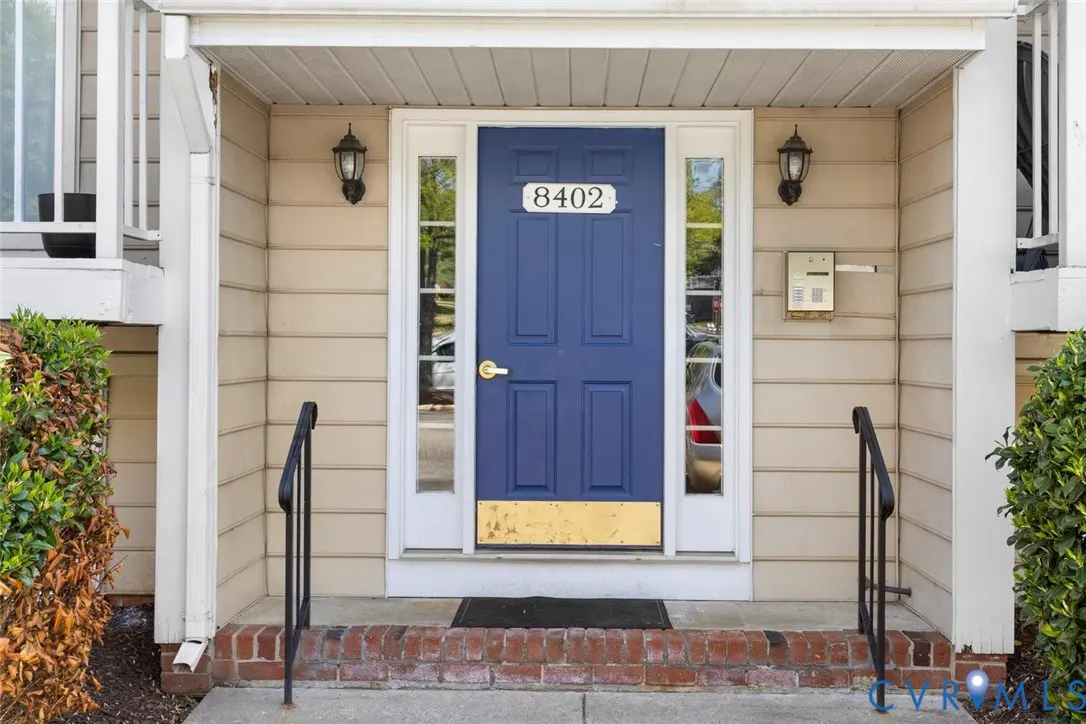 Front entrance of a home with a blue door, white trim, and the house number 8402 above the door. There are black handrails on either side of the brick steps.