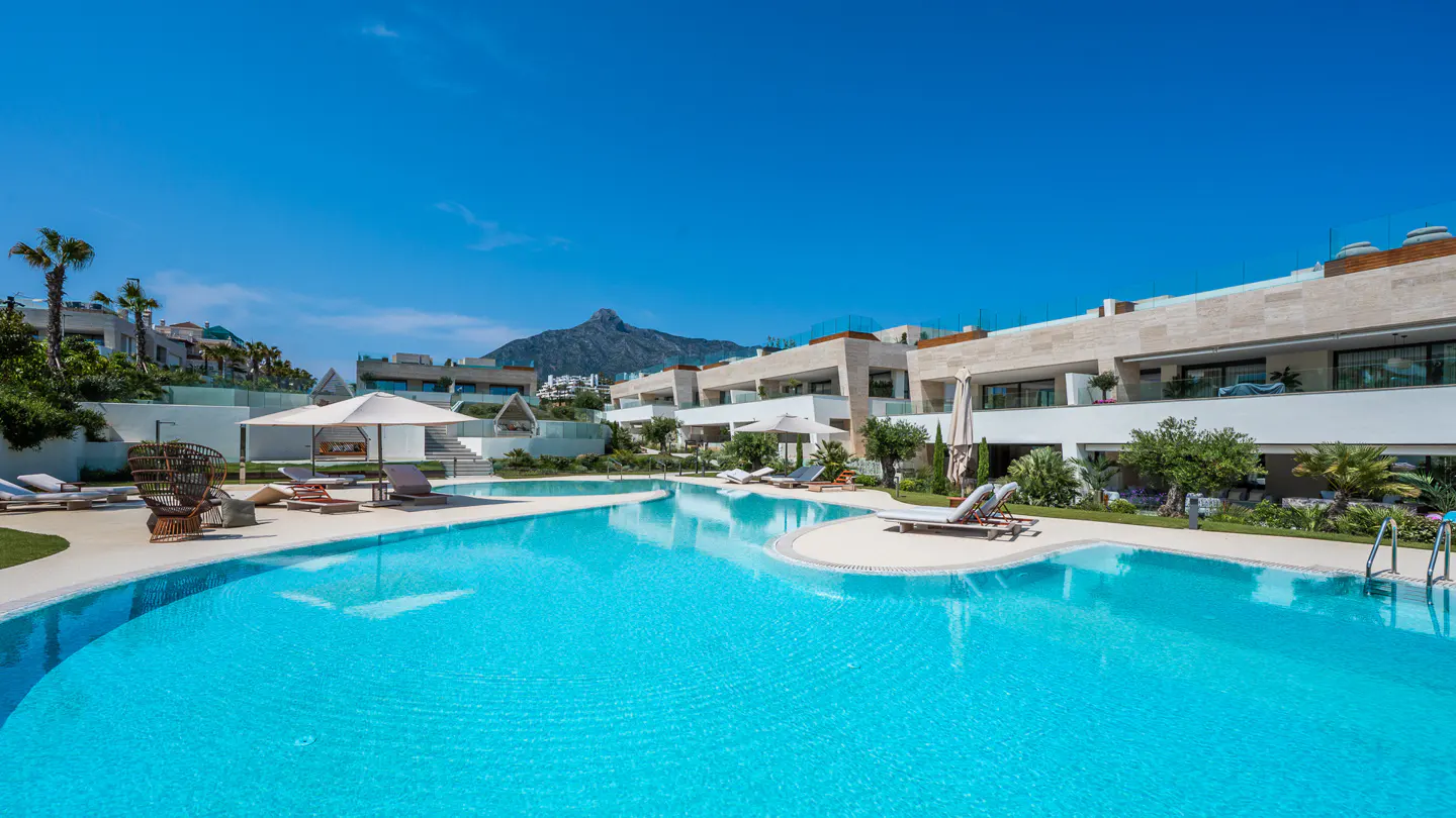 Luxury apartments surround a turquoise pool with lounge chairs and umbrellas under a clear blue sky. A mountain is visible in the background.