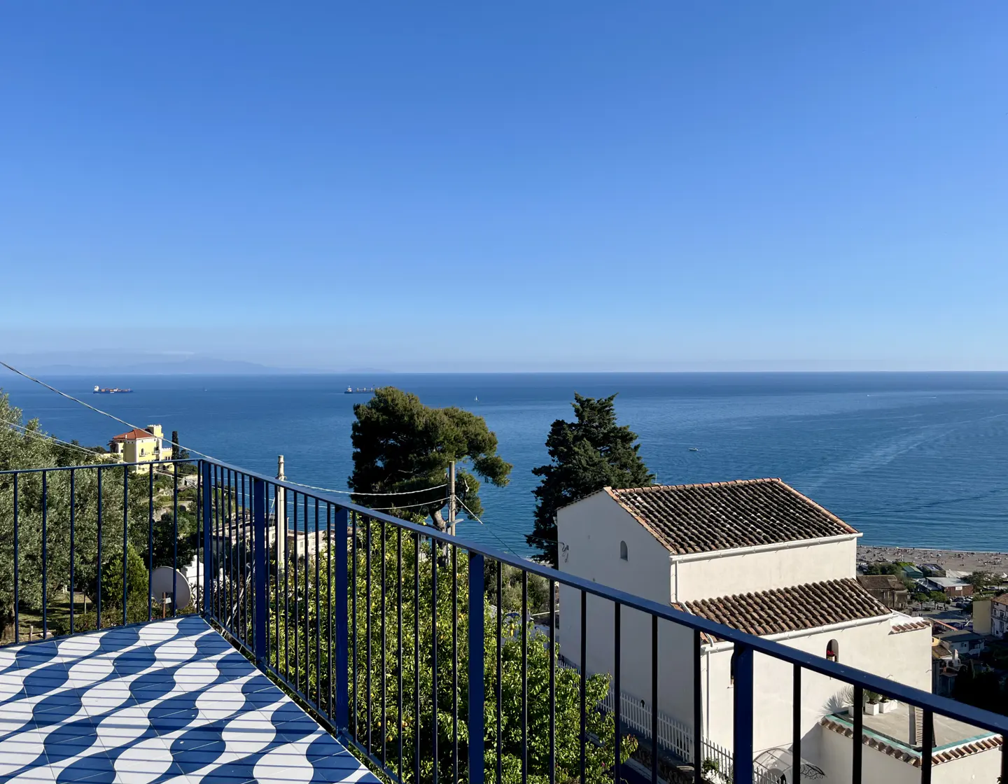 View from a blue-railed balcony with patterned tile, overlooking the ocean, trees, and a white building with a brown tile roof.