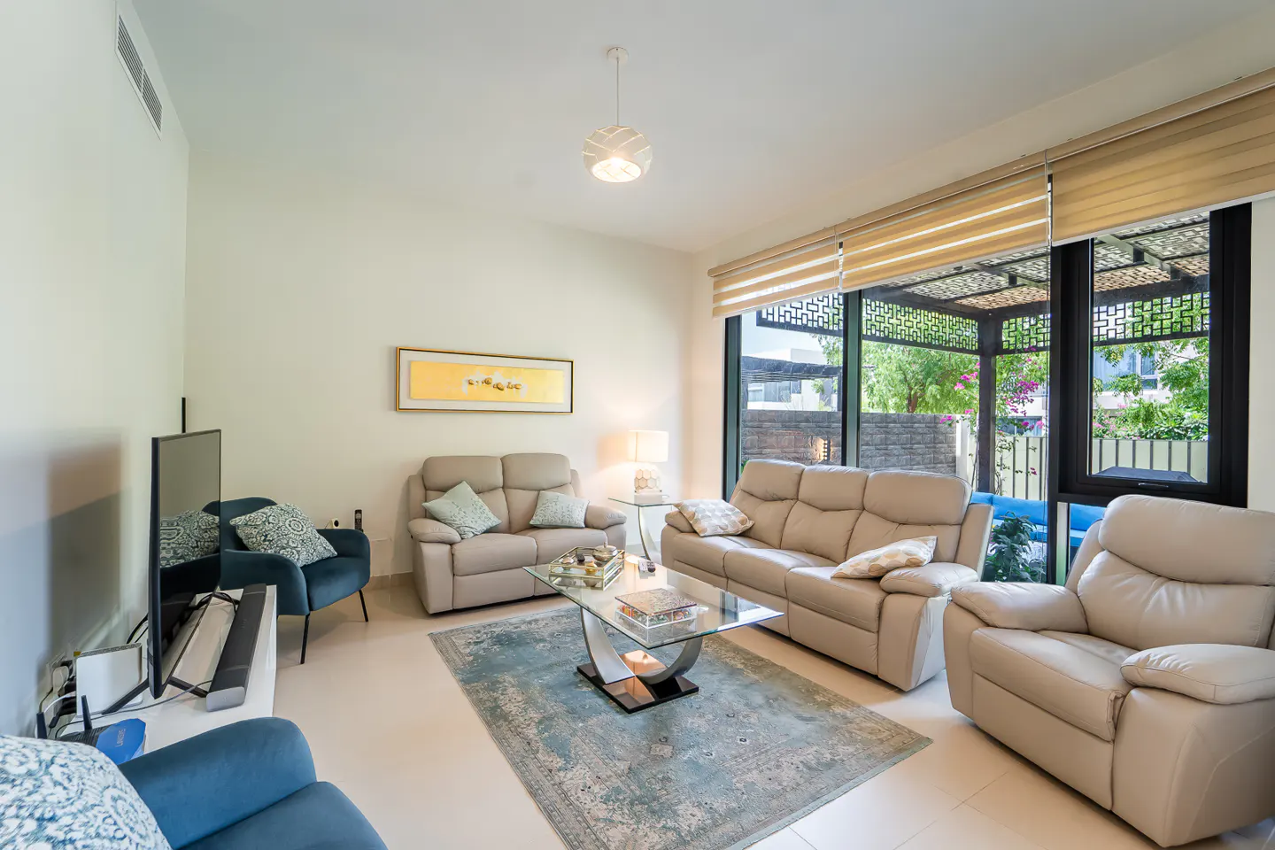 Living room with beige sofas, blue rug, glass table, and large windows overlooking a garden.