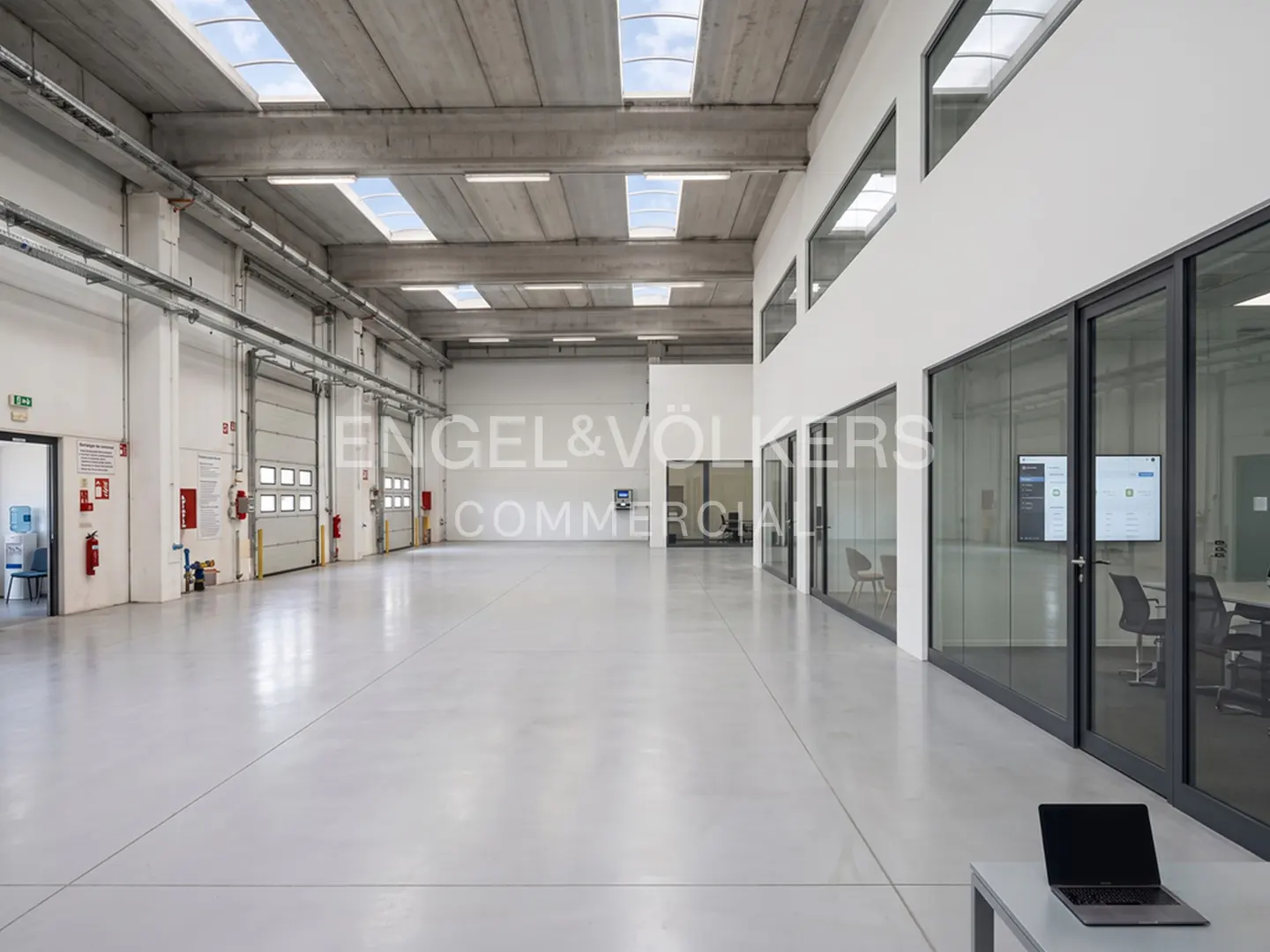 Bright, empty warehouse with white walls, gray floor, and skylights. Roll-up doors line one wall, and glass-walled offices are on the other.