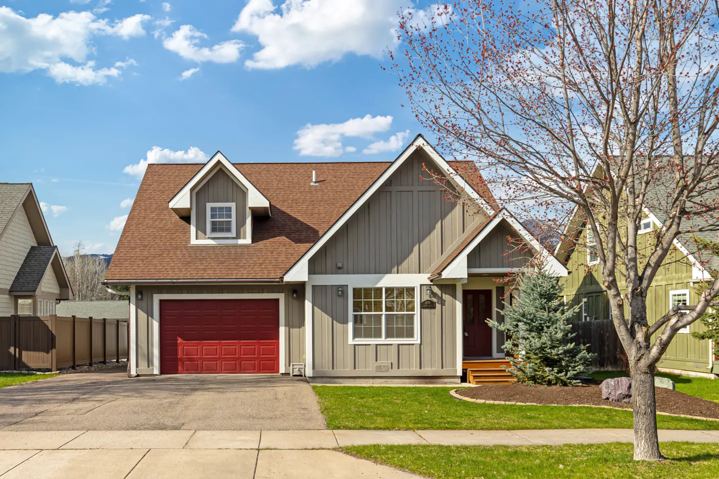 A gray, two-story house with a red garage door and brown roof on a sunny day.