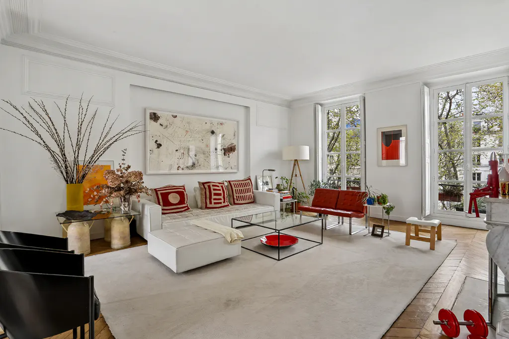 Bright living room with white walls, large windows, and herringbone wood floors. White sofa with red pillows, glass coffee table, and abstract art.
