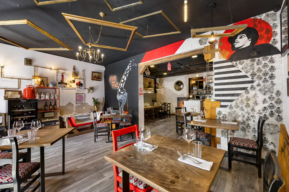 Restaurant interior with wooden tables, red chairs, and artistic wall murals. Empty gold picture frames hang from the black ceiling.