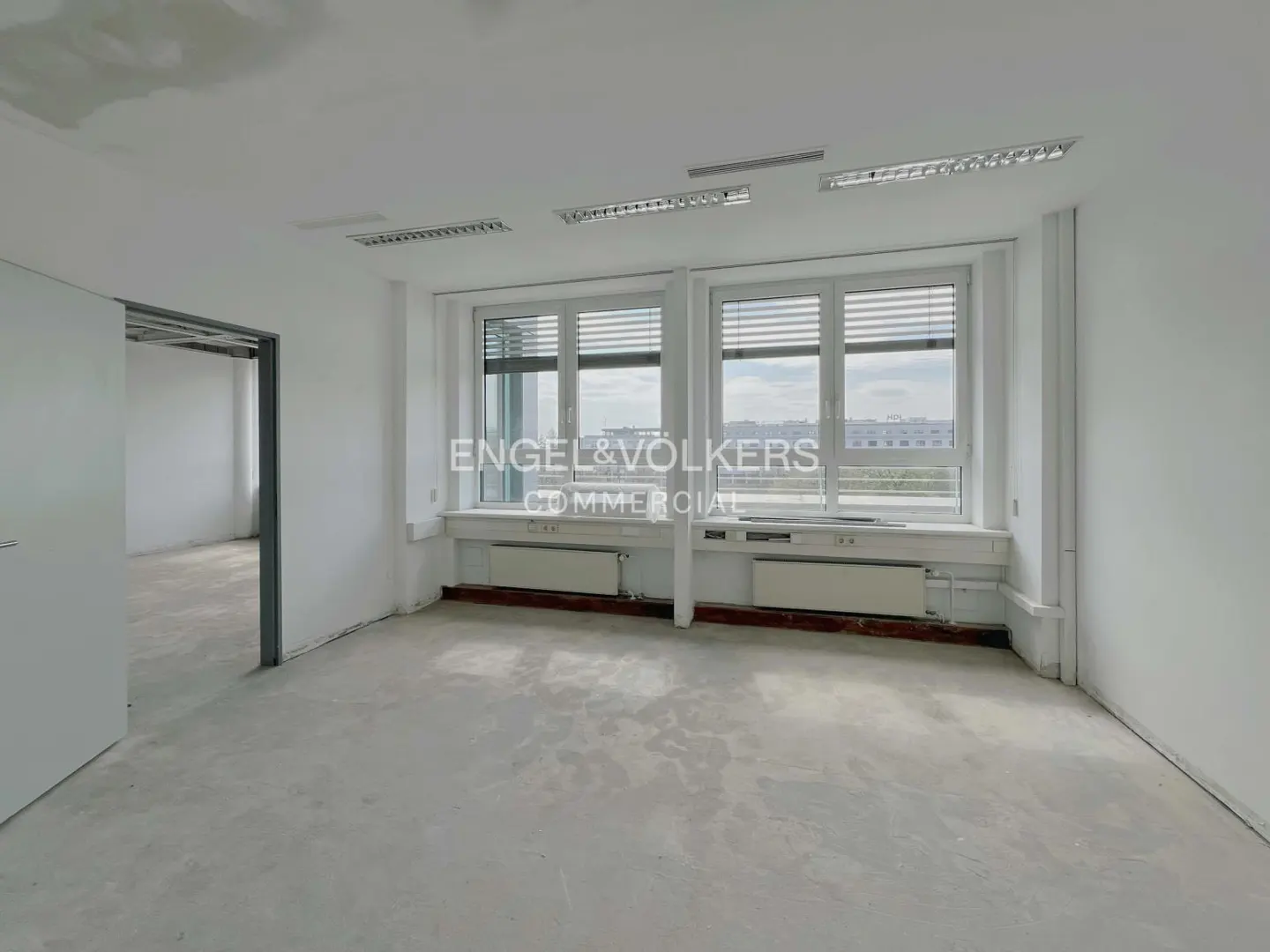 Empty white office space with large windows, radiators, and fluorescent lighting. Doorway to another room is on the left.