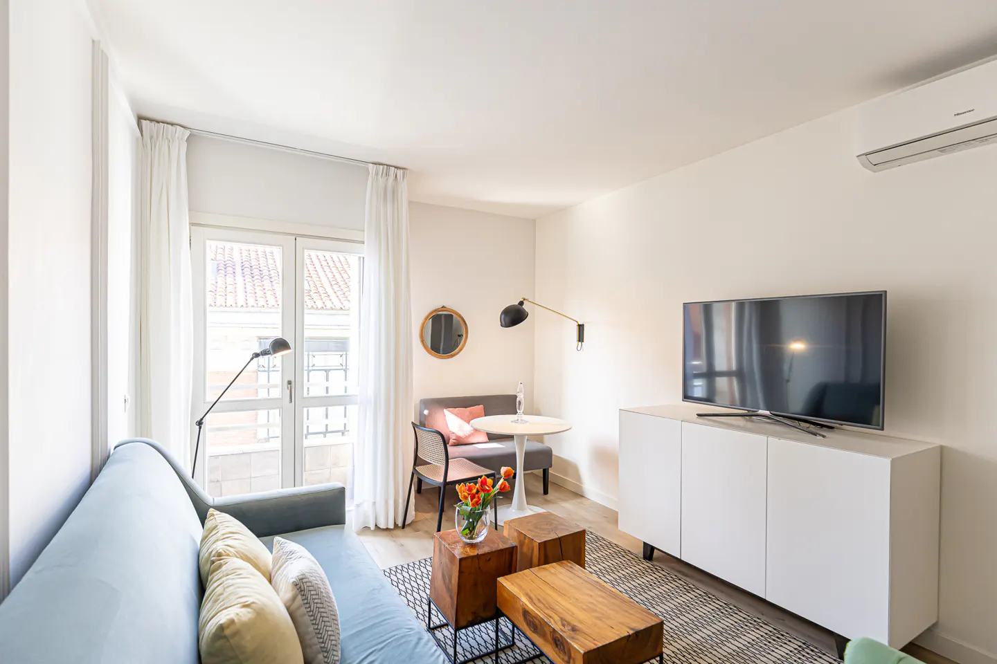 Bright living room with a blue sofa, wooden coffee tables, and a white media console with a TV. A small dining area is near a window.