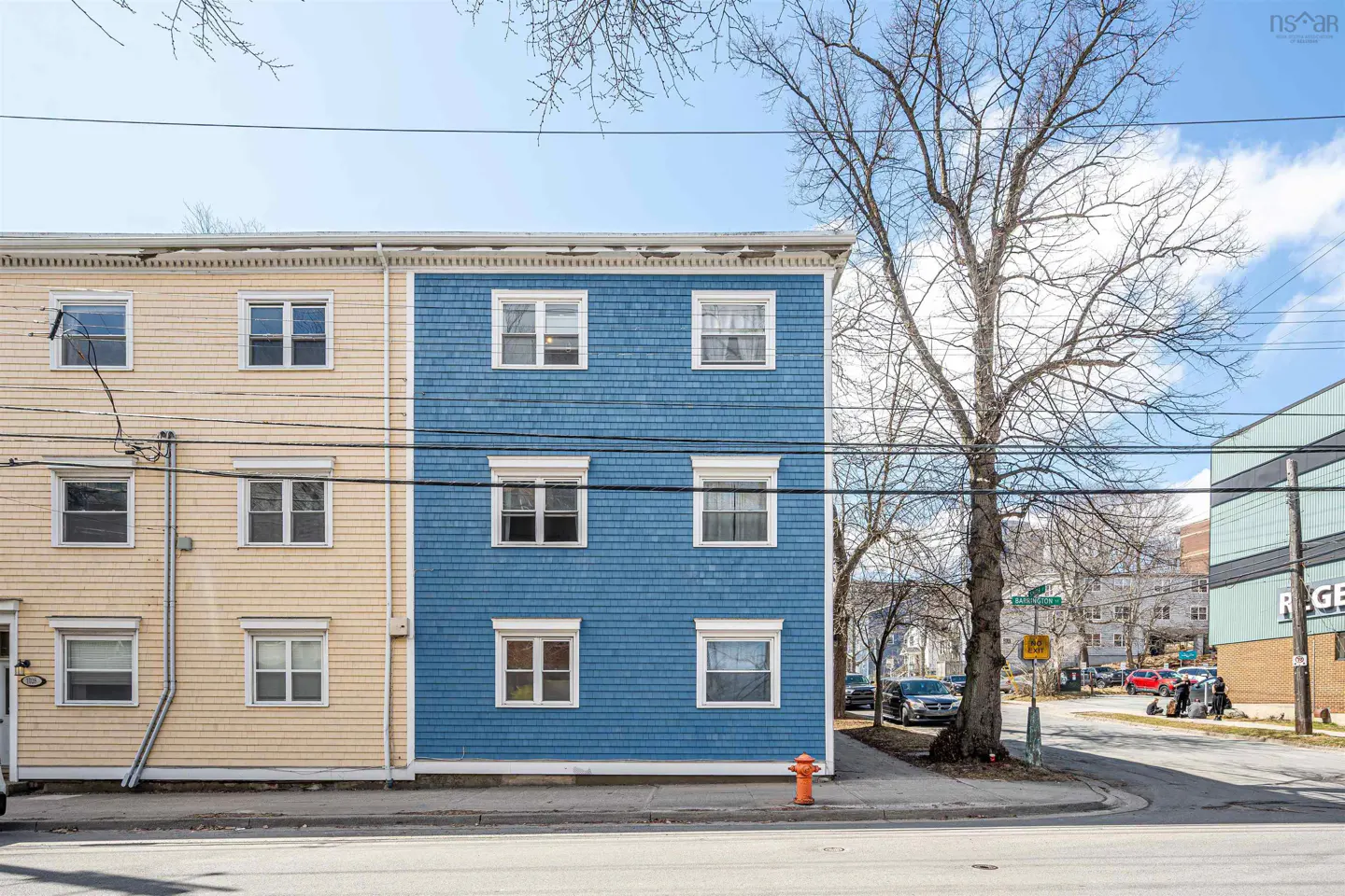 Exterior view of a three-story building with blue and yellow siding and white trim. A tree stands to the right of the building.