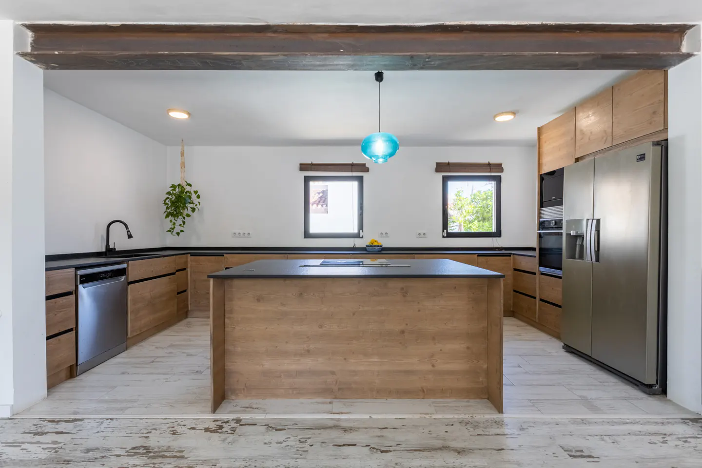A bright kitchen with wood cabinets, black countertops, and a large island. A blue pendant light hangs above the island. Stainless steel appliances are visible.