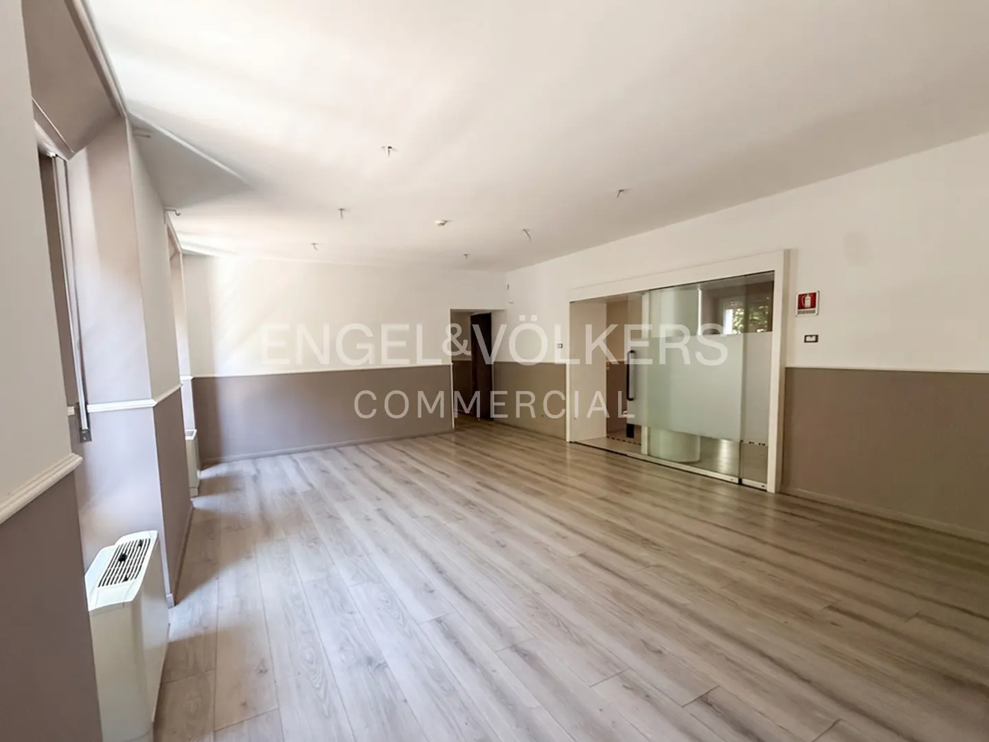 Empty commercial office space with light wood-look flooring, white and beige walls, and a glass-partitioned room.