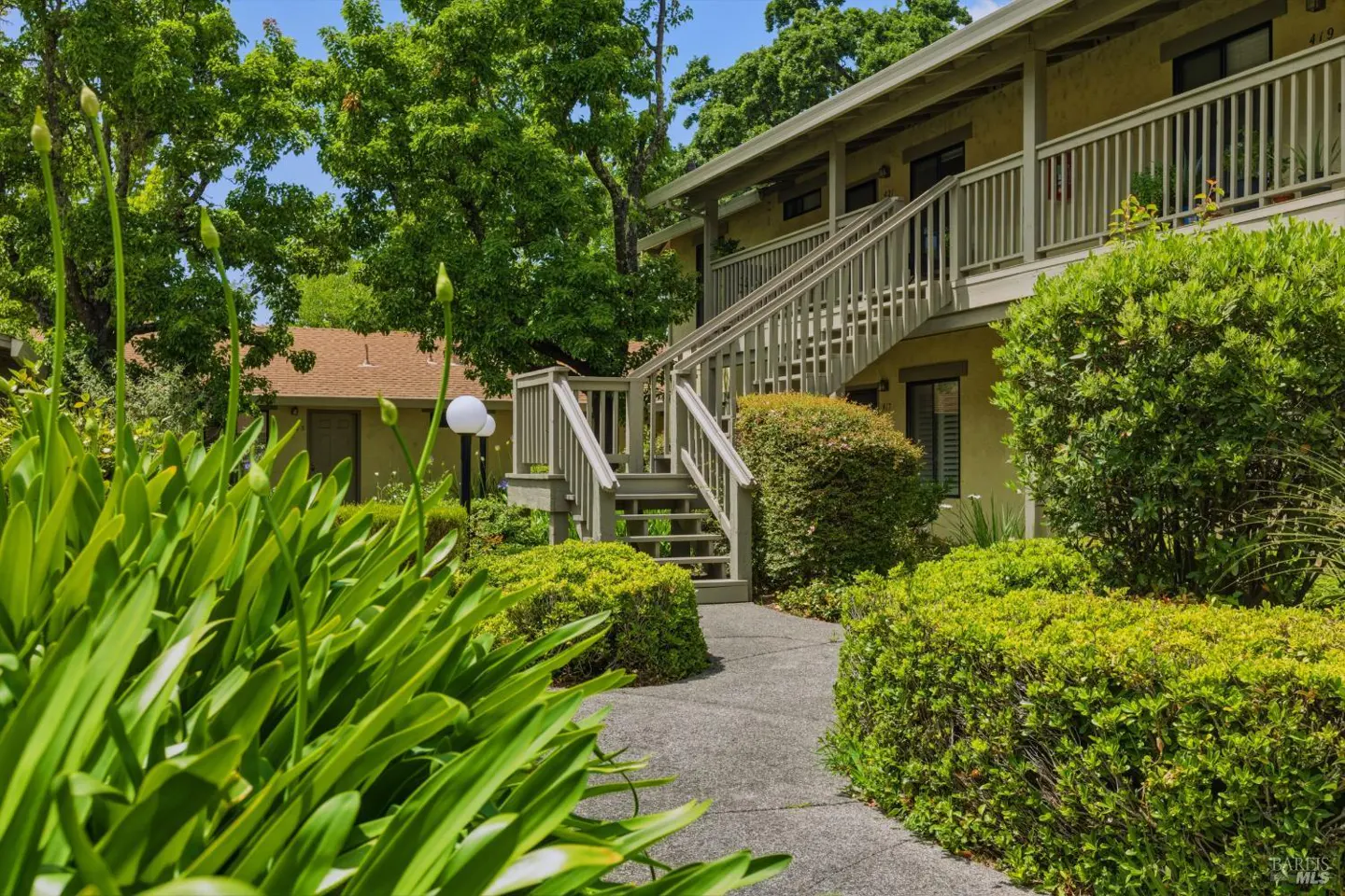 A concrete path leads to a wooden staircase outside a two-story tan apartment building surrounded by lush green landscaping.