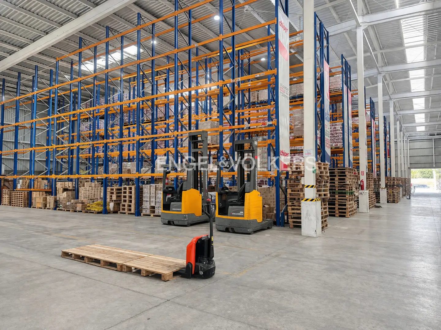 A large warehouse interior with blue and orange shelving, forklifts, and stacks of wooden pallets.