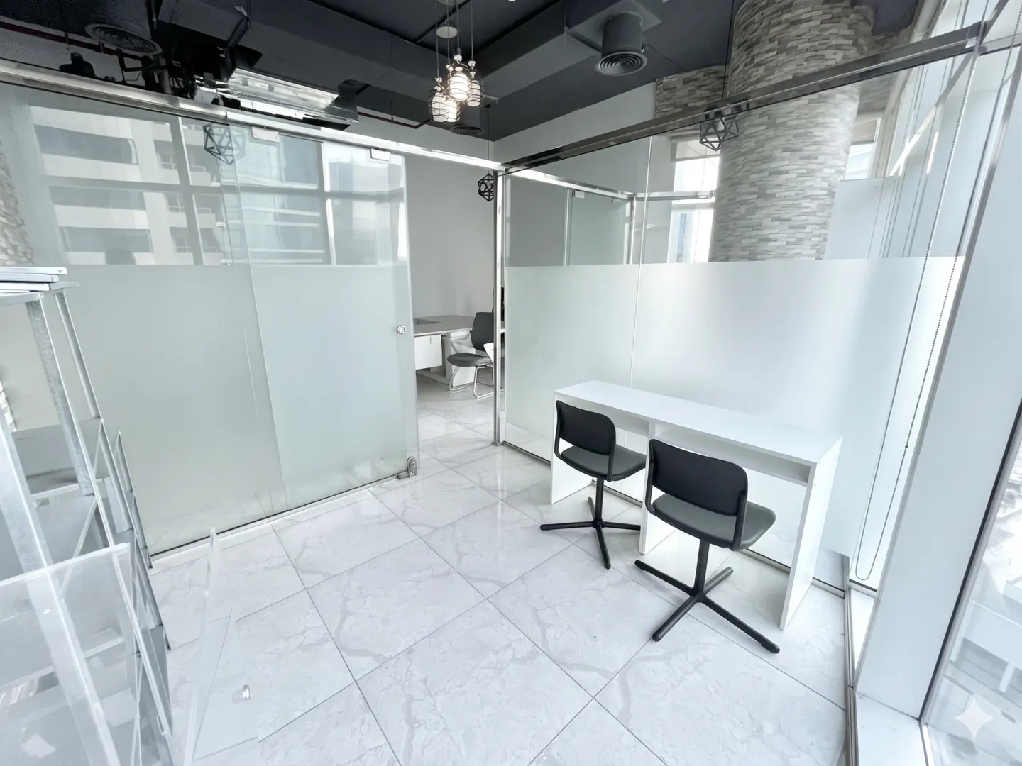 Bright office space with white tile floors, glass-walled rooms, white desks, and black chairs.