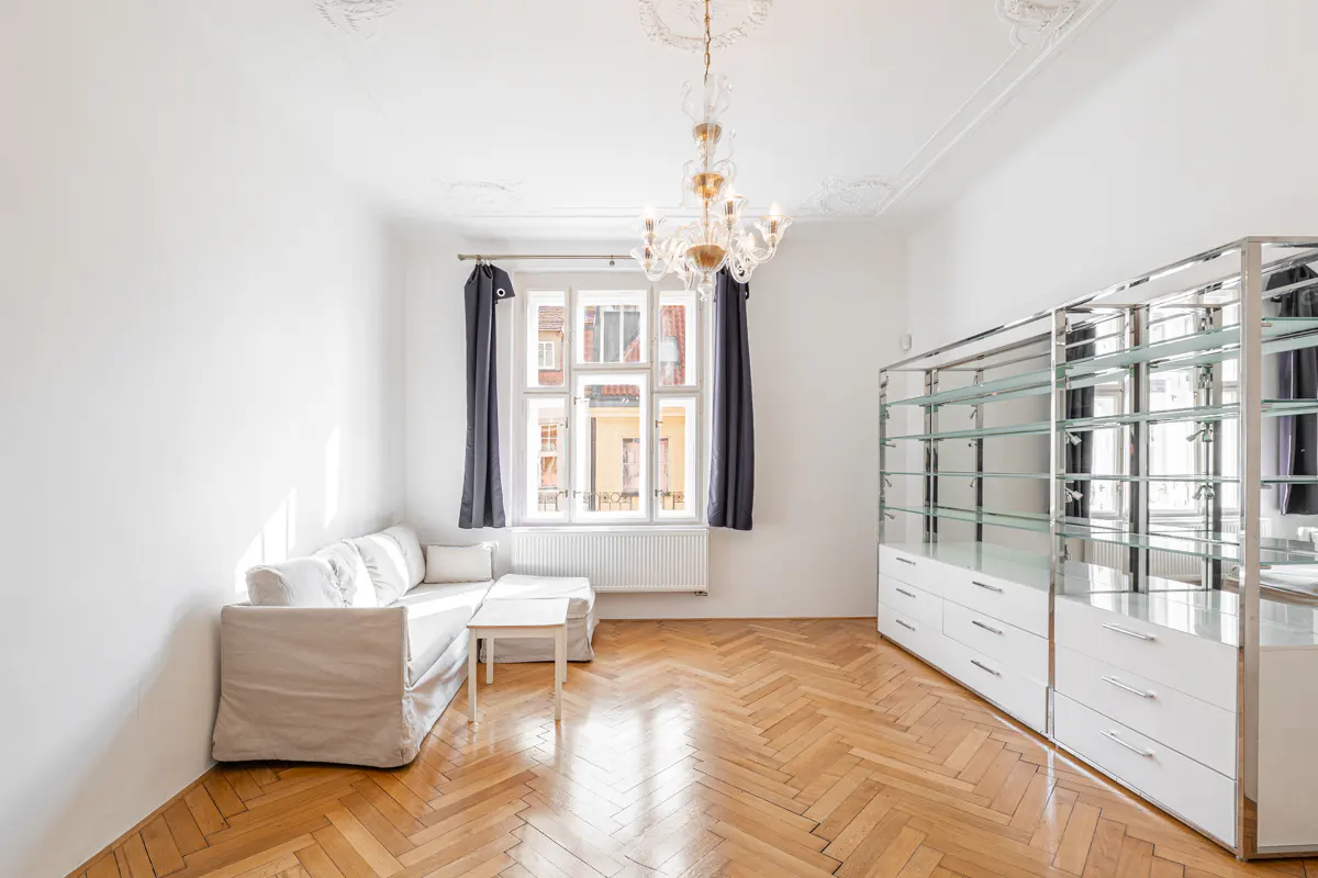 Bright living room with herringbone wood floors, white walls, and a crystal chandelier. A white sofa sits near a window with dark curtains.
