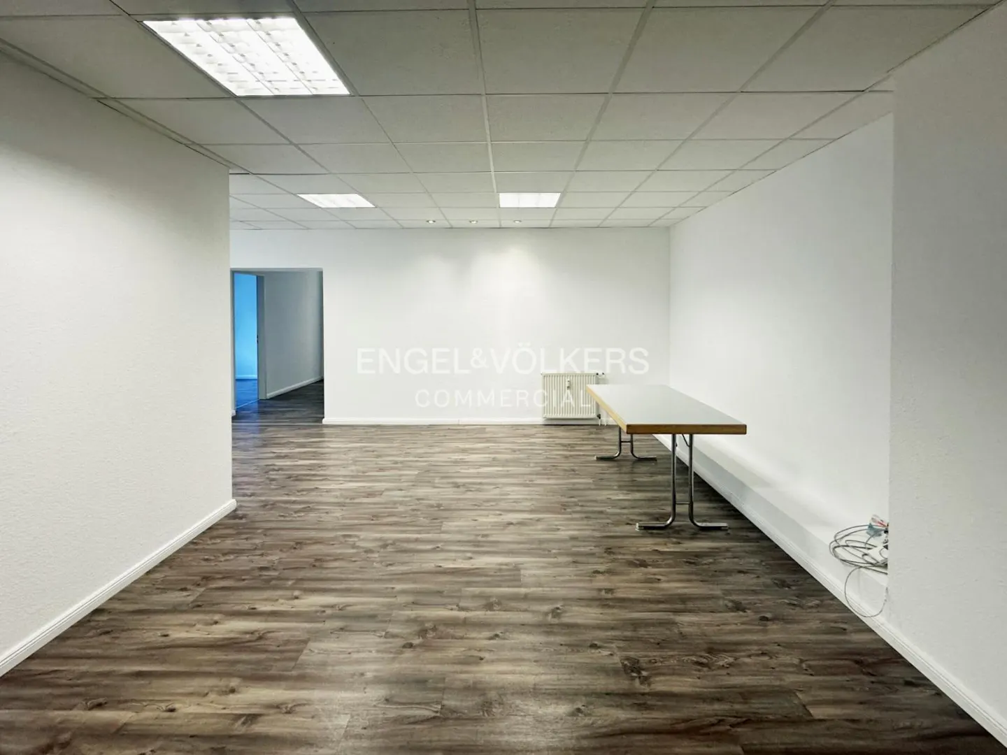 Empty commercial space with wood-look floors, white walls, and a drop ceiling with fluorescent lights. A table sits against the wall.