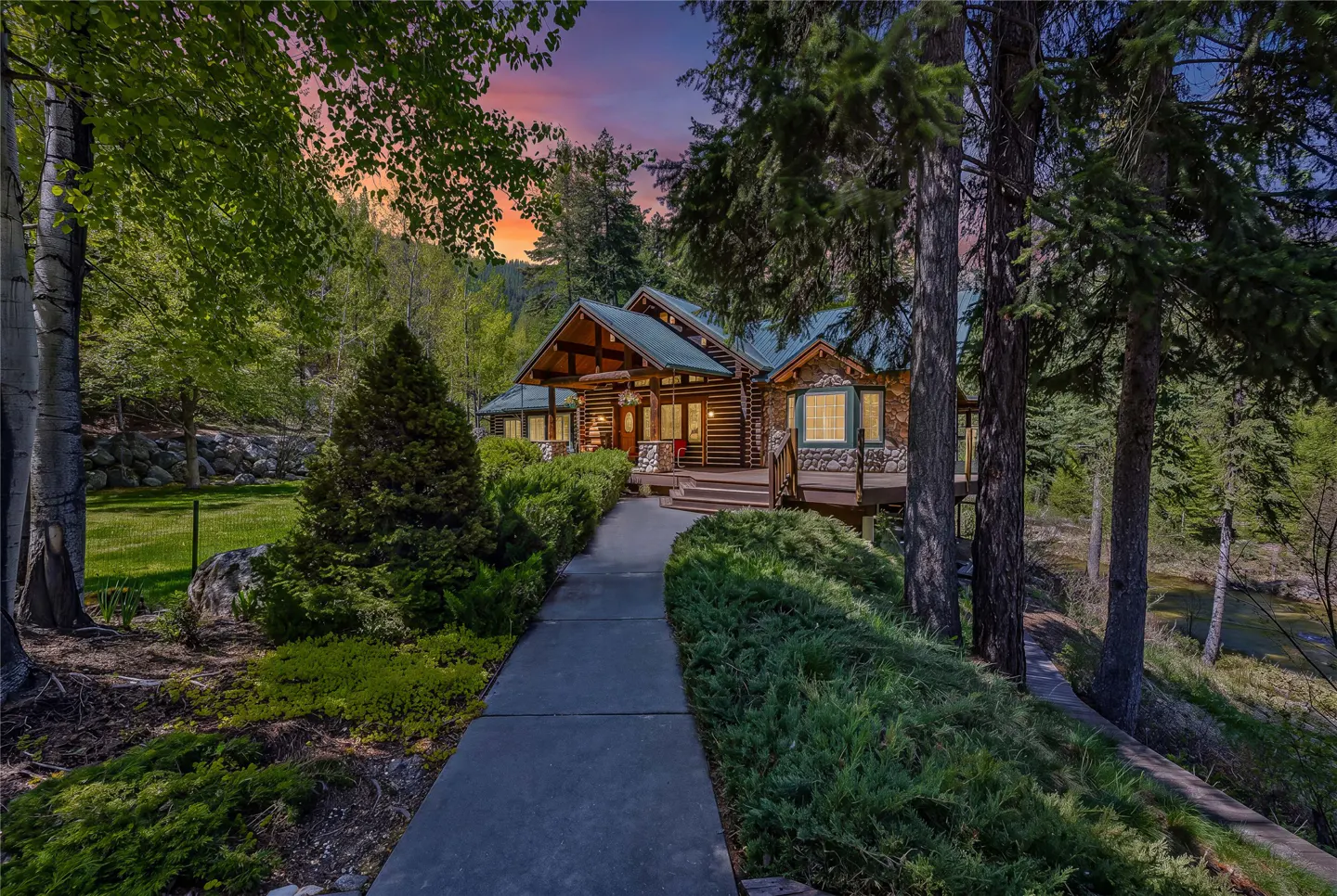 Log cabin home with a green roof, stone accents, and a walkway leading to the front door, surrounded by lush greenery and trees.