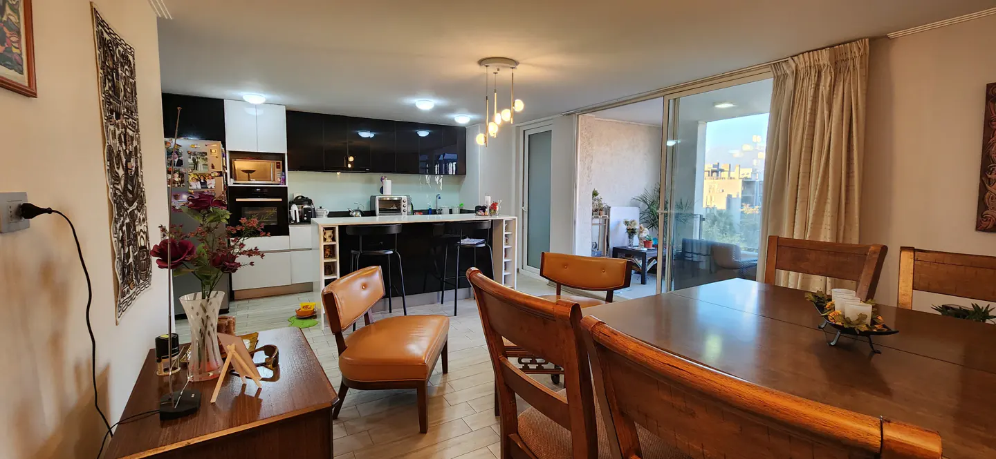 A bright, modern apartment interior with a dining table, chairs, and a kitchen with black and white cabinets. A balcony is visible through sliding glass doors.