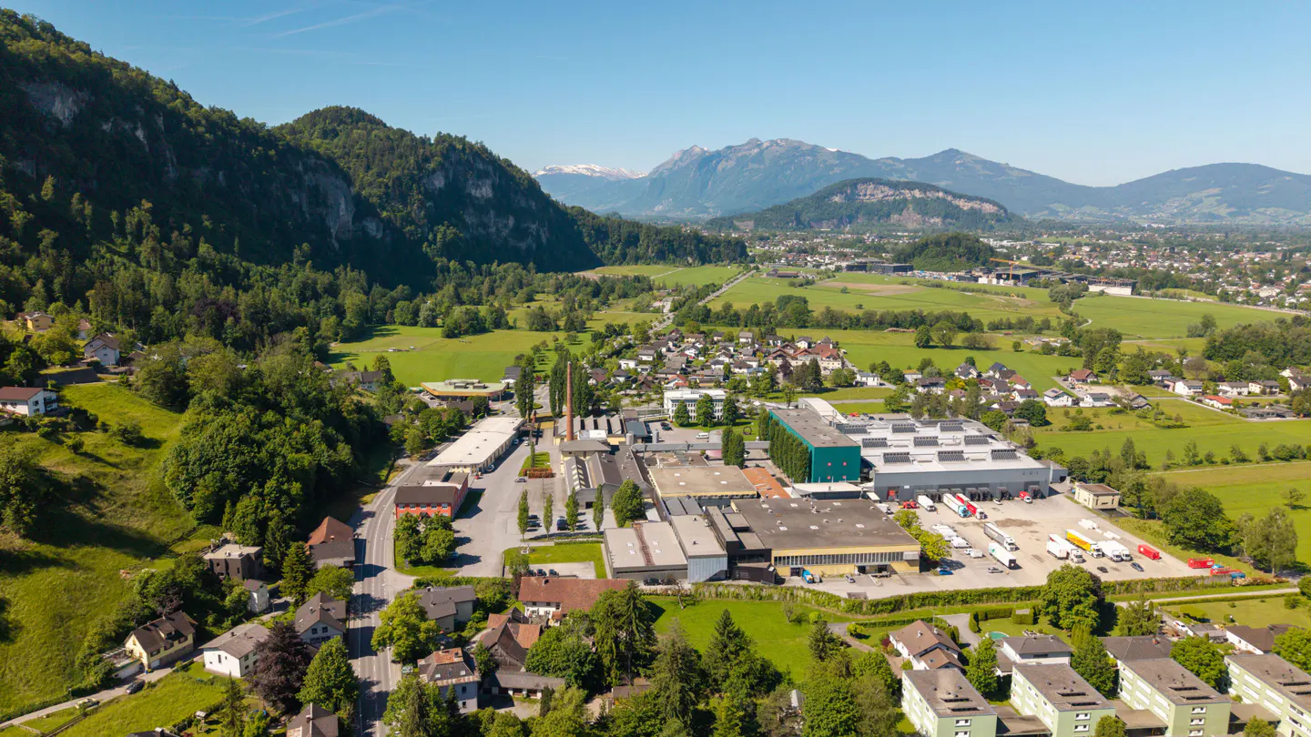 Aerial view of a factory complex nestled in a valley, surrounded by green hills and distant mountains under a clear blue sky.