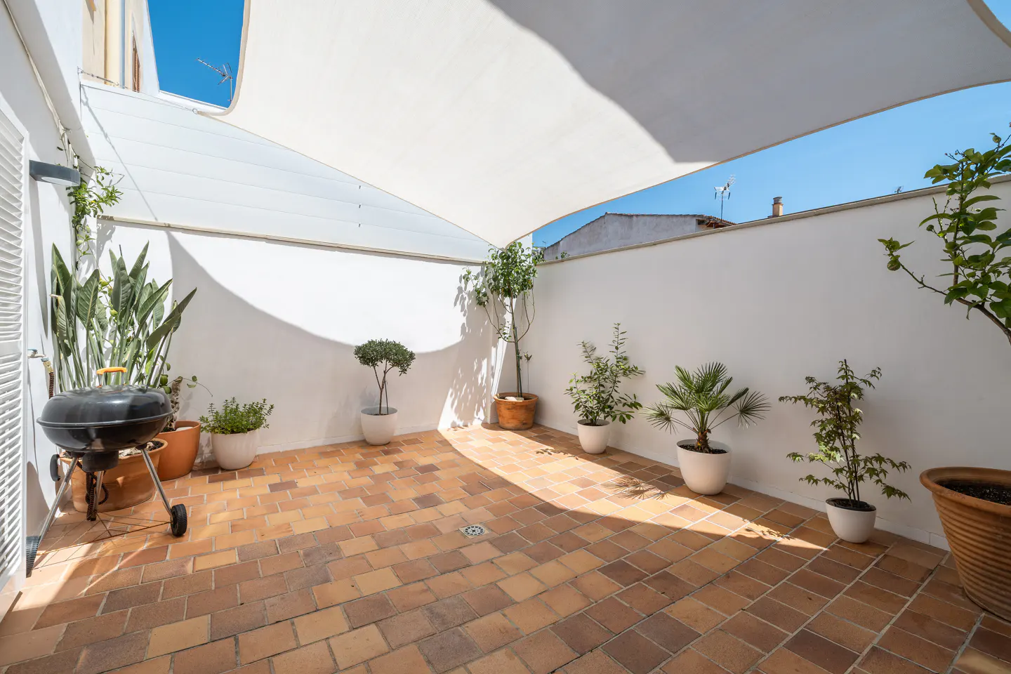 Outdoor patio with brick flooring, white walls, and a white shade sail overhead. Potted plants and a black grill add to the cozy atmosphere.