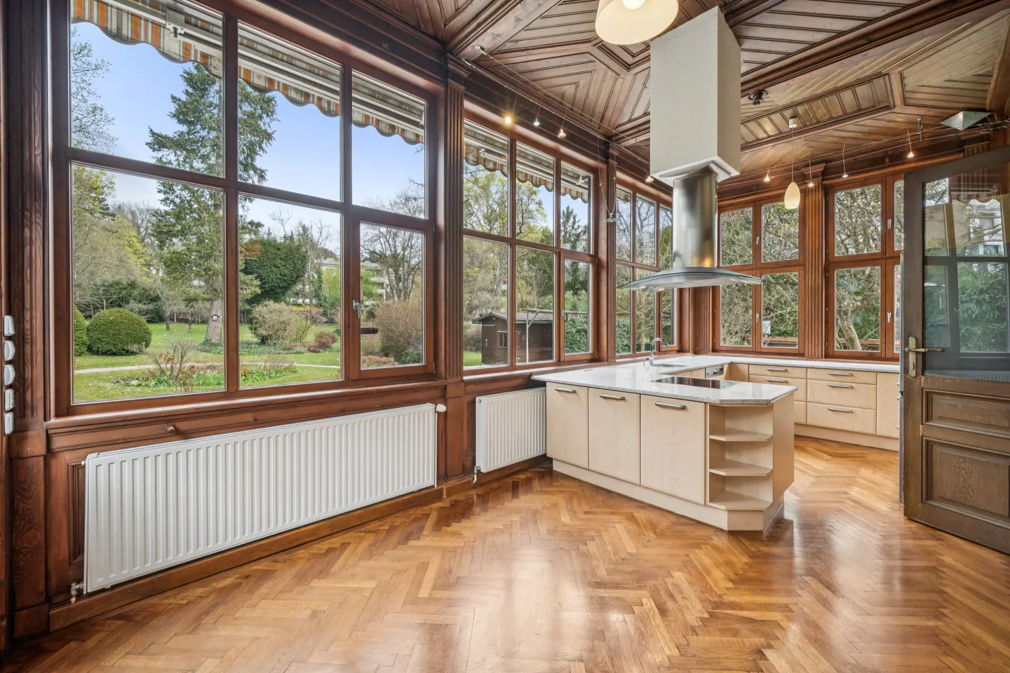 A bright kitchen with wood floors, cabinets, and trim. Large windows look out onto a green garden.