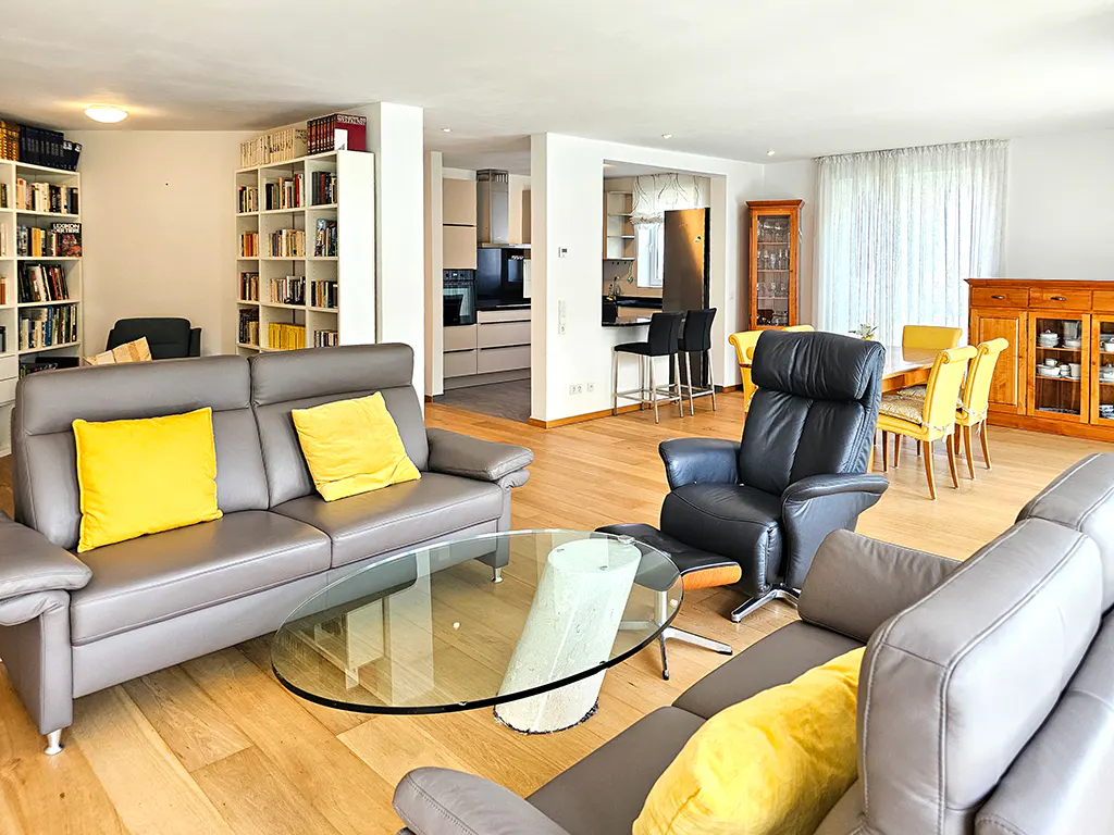A bright living room with gray sofas, yellow pillows, a glass table, and a black recliner. Bookshelves and a kitchen are in the background.