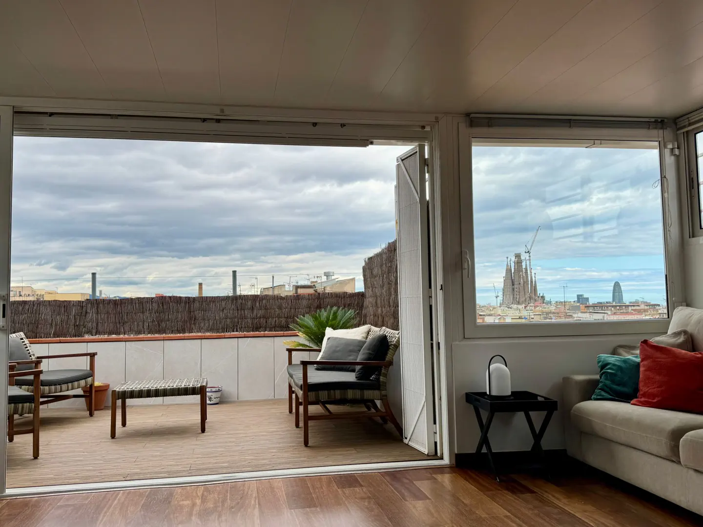 Indoor view of a Barcelona apartment with a balcony. The Sagrada Familia is visible through the window. The balcony has chairs and a table.