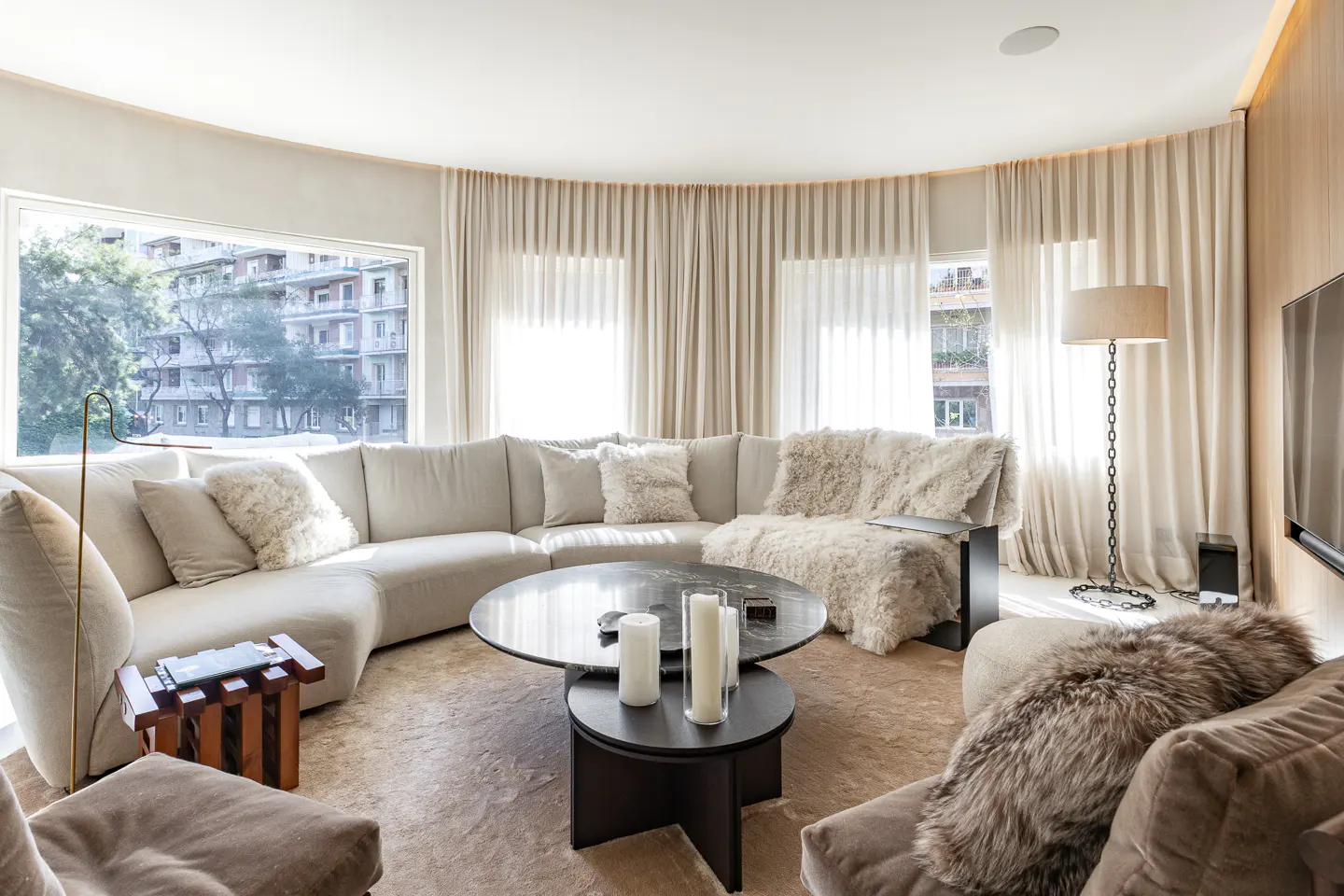 Modern living room with a curved cream sofa, round marble coffee table, plush fur throws, and floor-to-ceiling curtains.