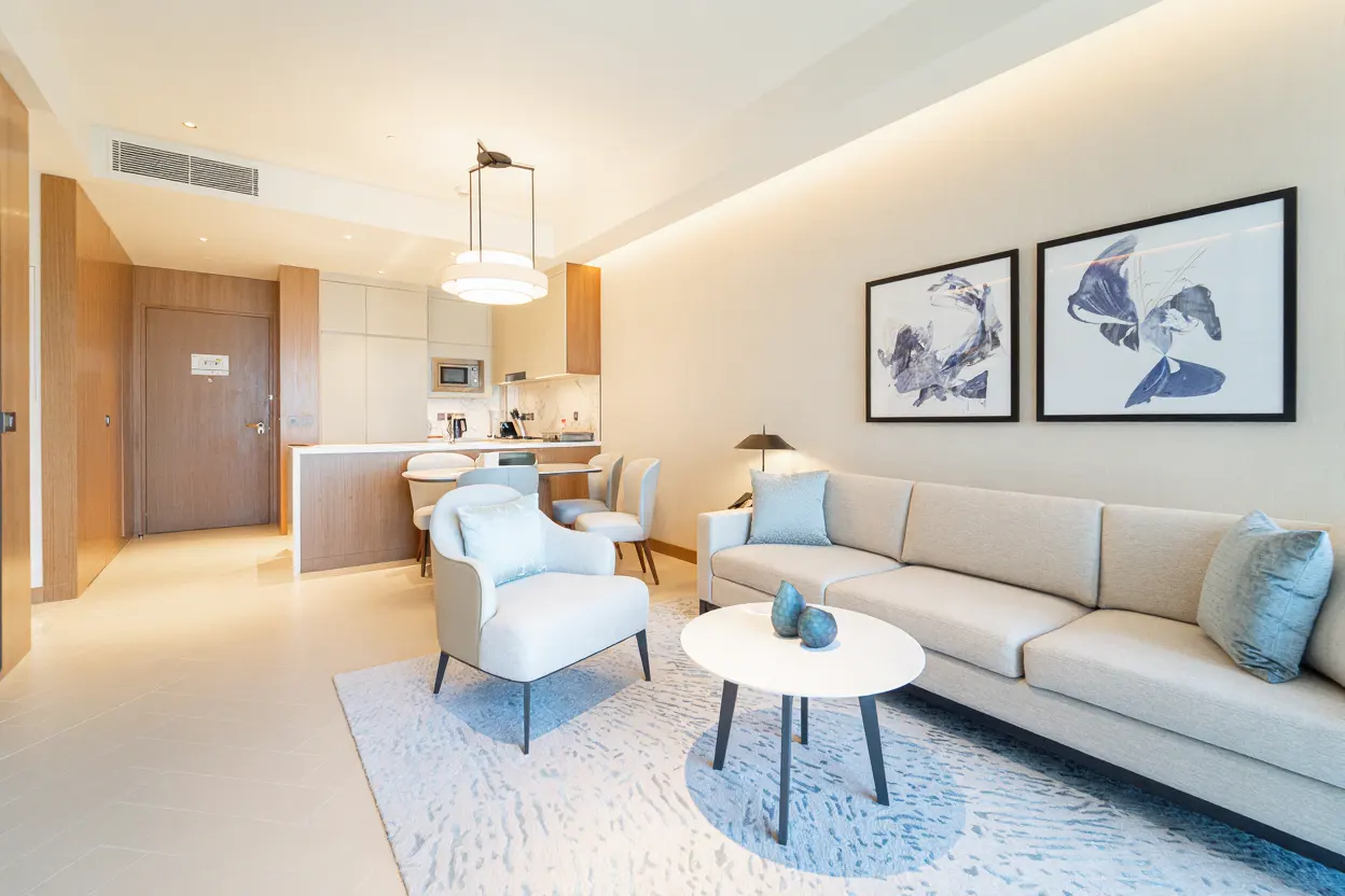 A bright, modern apartment interior with a sofa, chair, and kitchen area. Two framed art pieces hang above the sofa.