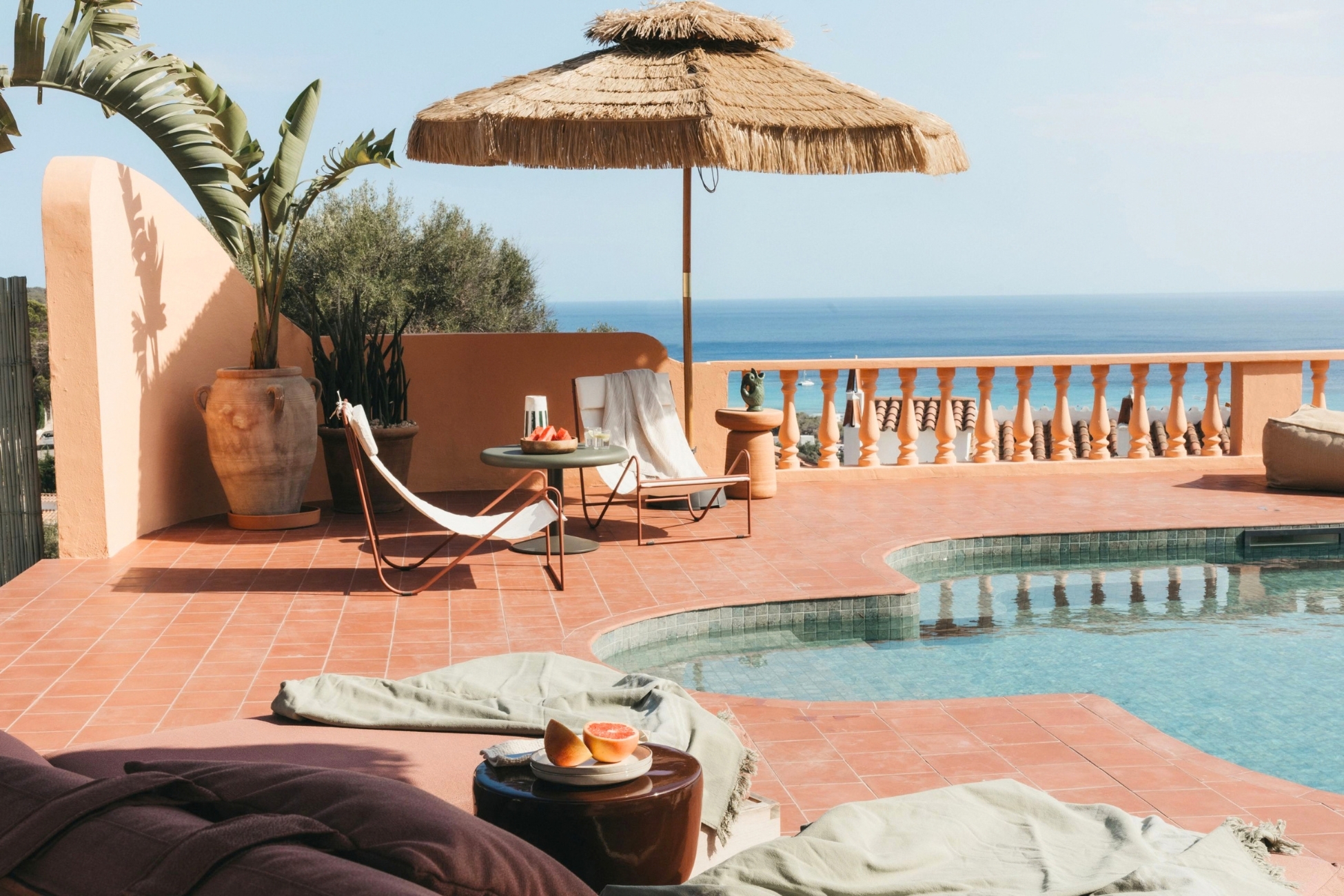 Terracotta patio with a swimming pool, lounge chairs, a straw umbrella, and a view of the blue ocean under a clear sky. [AI-generated text]