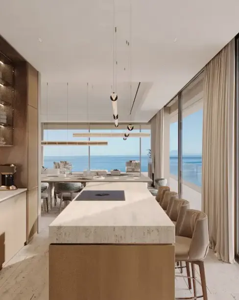 A modern kitchen with a large island, beige chairs, and ocean view through large windows.