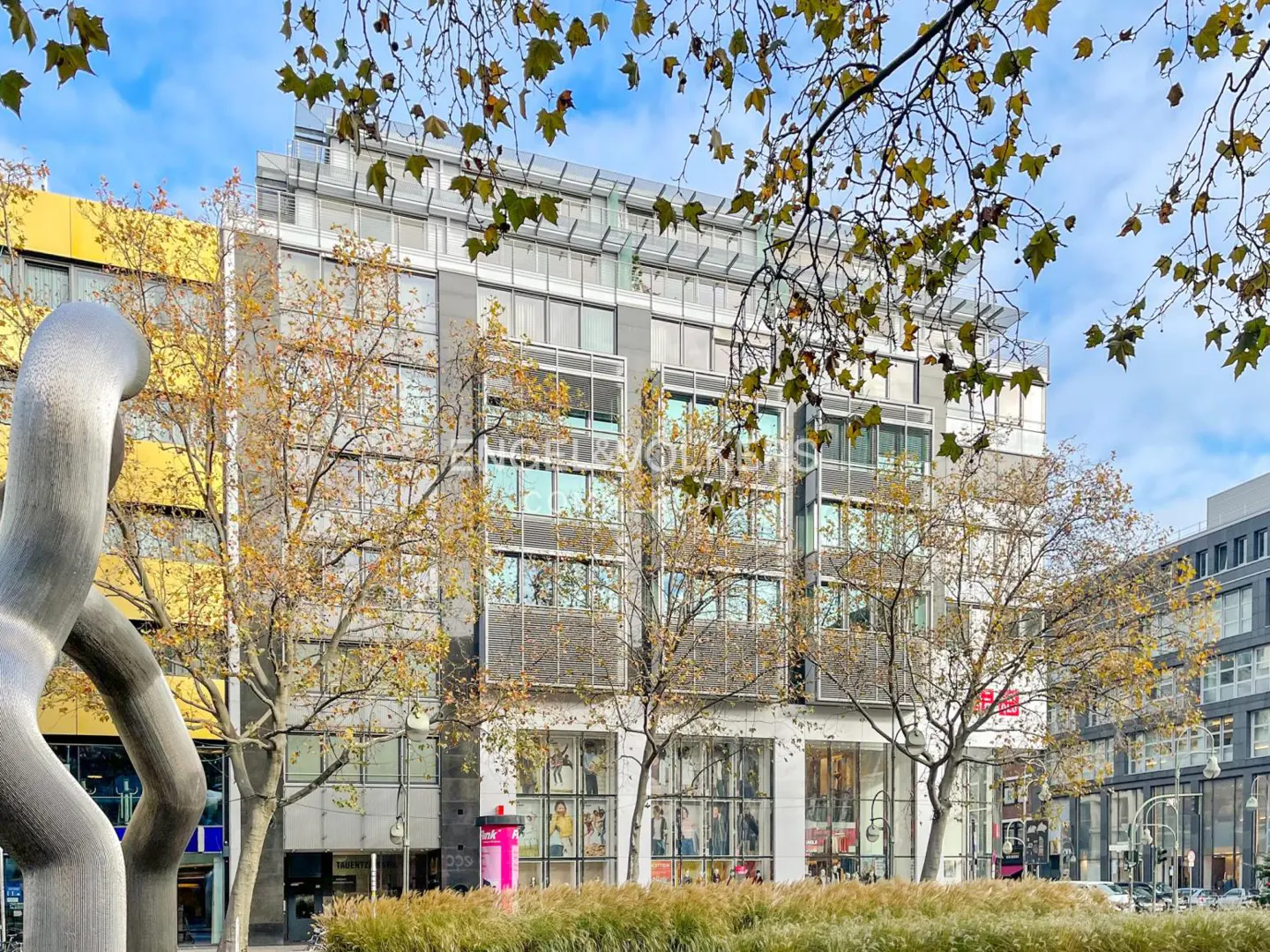 Modern building with large windows and a gray facade, framed by trees with autumn leaves and a silver sculpture.
