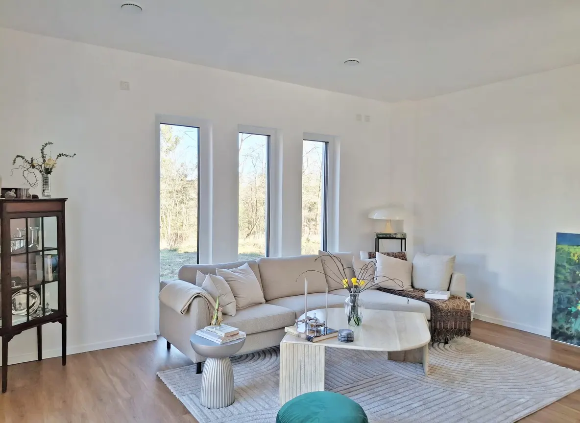 Bright living room with white walls, wood floors, and three tall windows. A beige sectional sofa sits on a patterned rug, with a marble coffee table and a green ottoman.