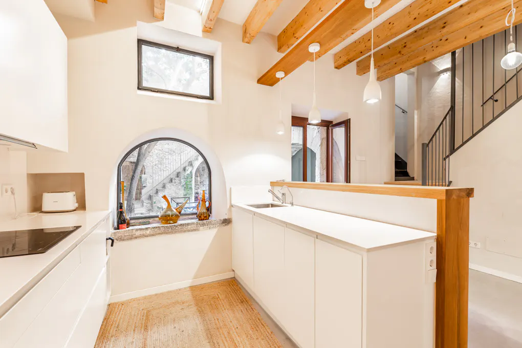 Bright kitchen with white cabinets, wooden beams, and arched window. A staircase is visible in the background.