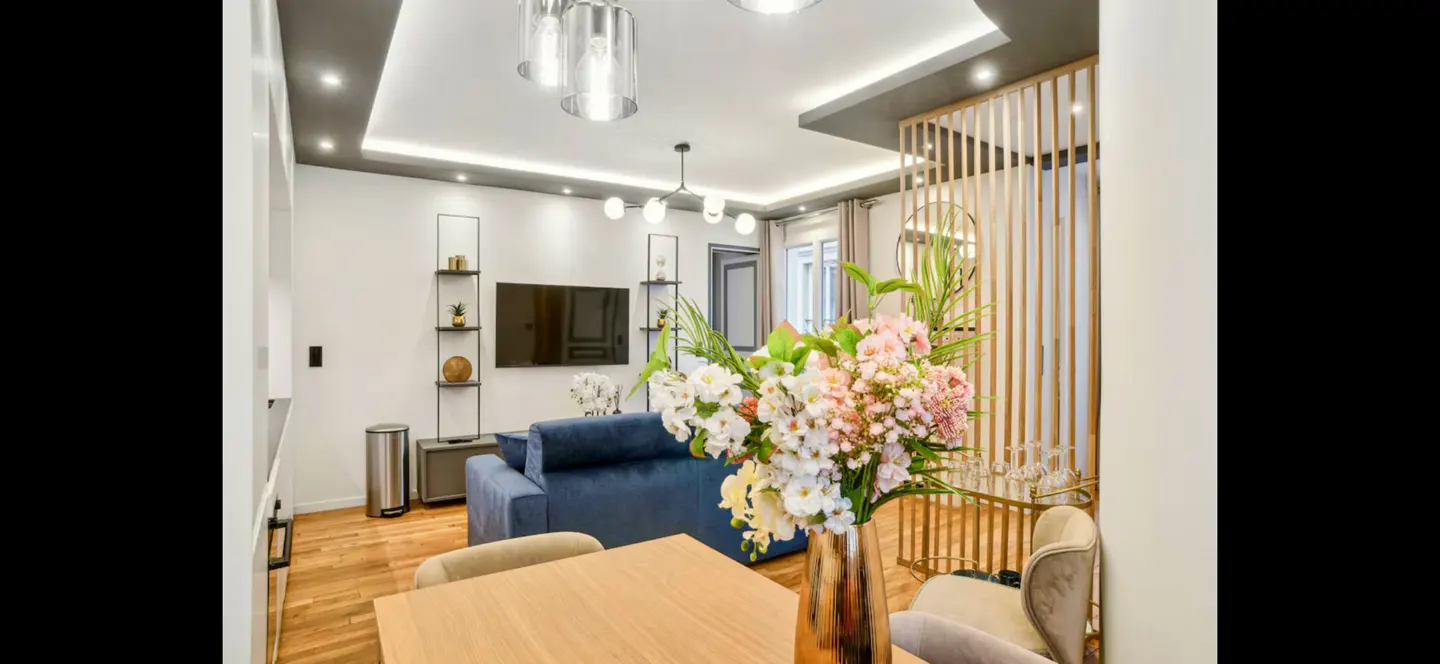 Bright living room with a blue sofa, wood floors, and a table with a vase of pink and white flowers.