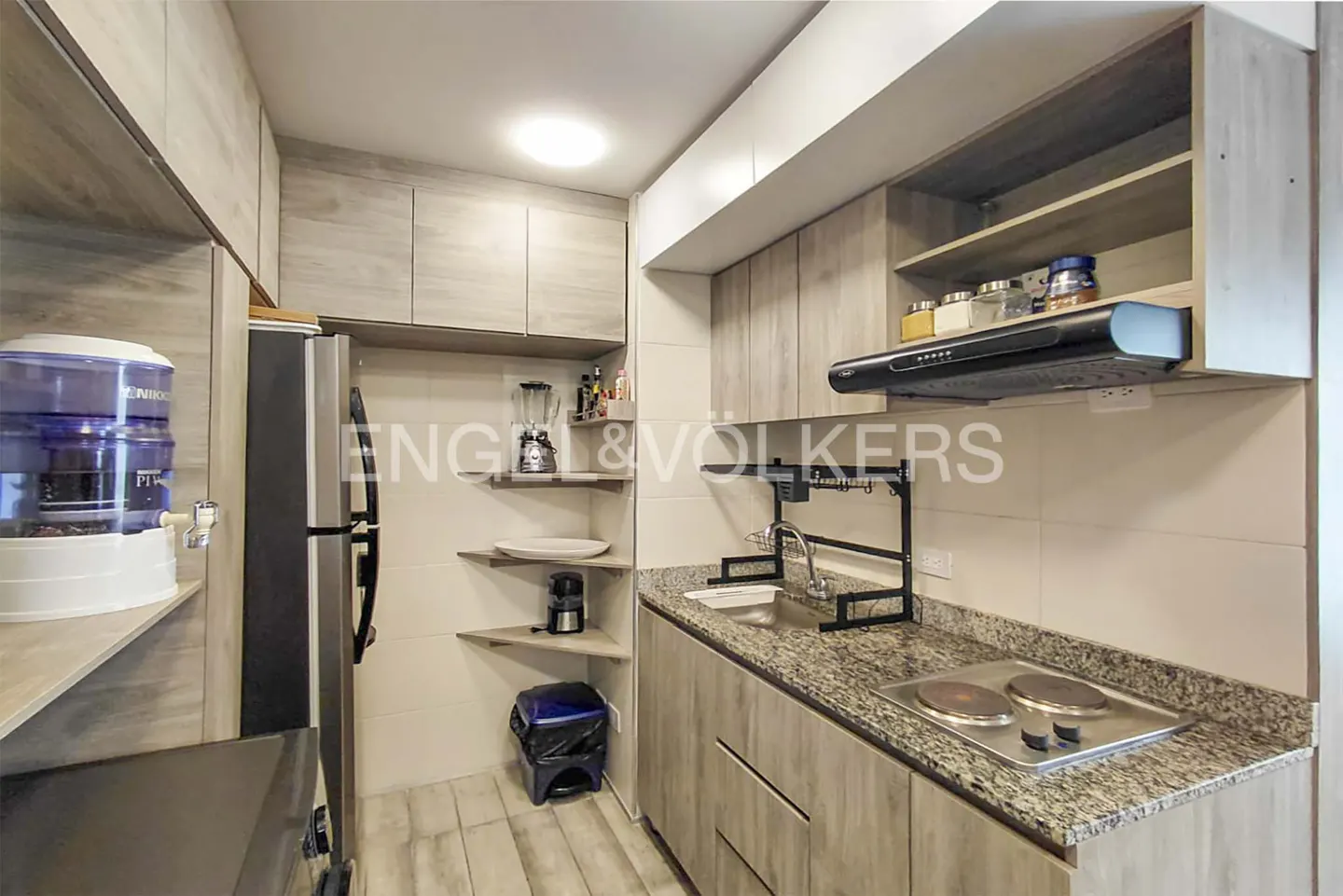 A compact kitchen with light wood cabinets, granite countertops, and stainless steel appliances. Shelves hold a blender and coffee maker.