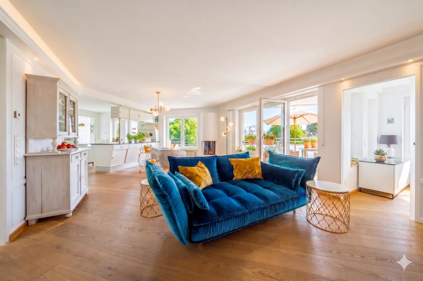 Bright open-plan living room with a plush blue velvet sofa, gold side tables, and a view of a sunny balcony.