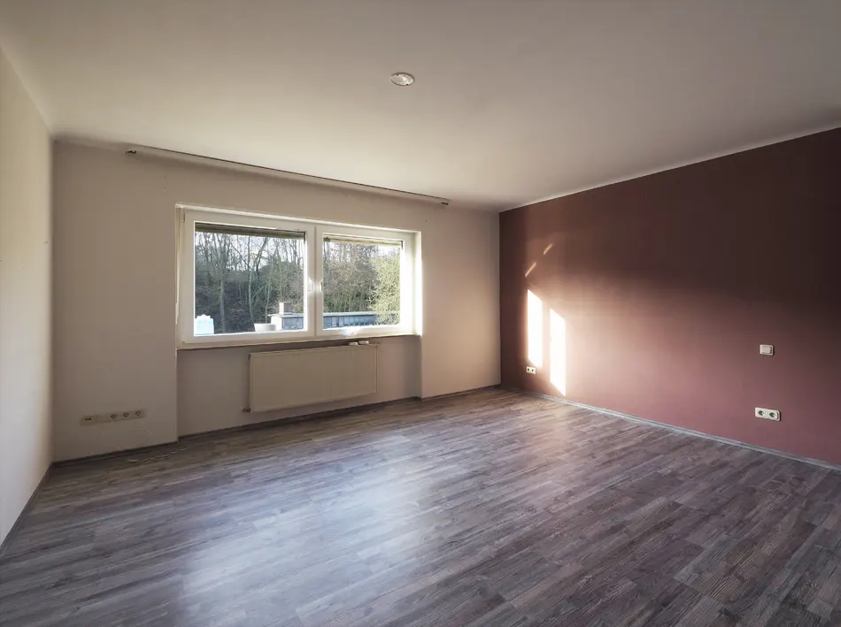 Empty room with wood-look floors, white walls, and a maroon accent wall. A window lets in sunlight.